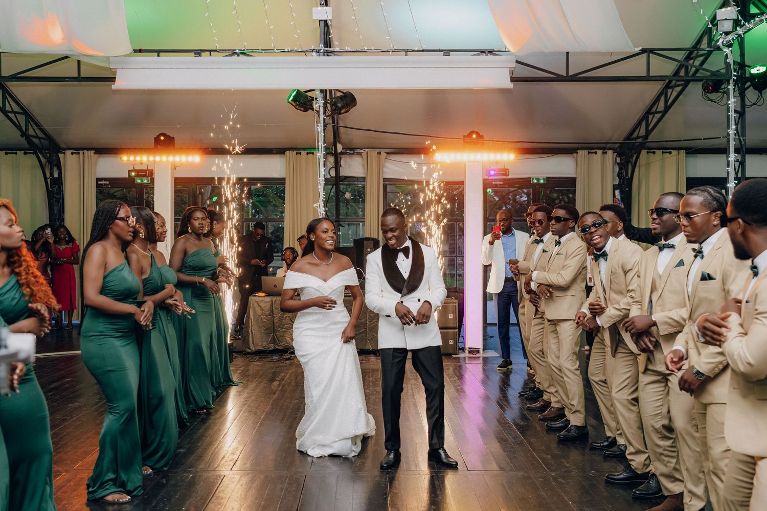 Wedding party in green and beige attire dancing together during reception celebration.