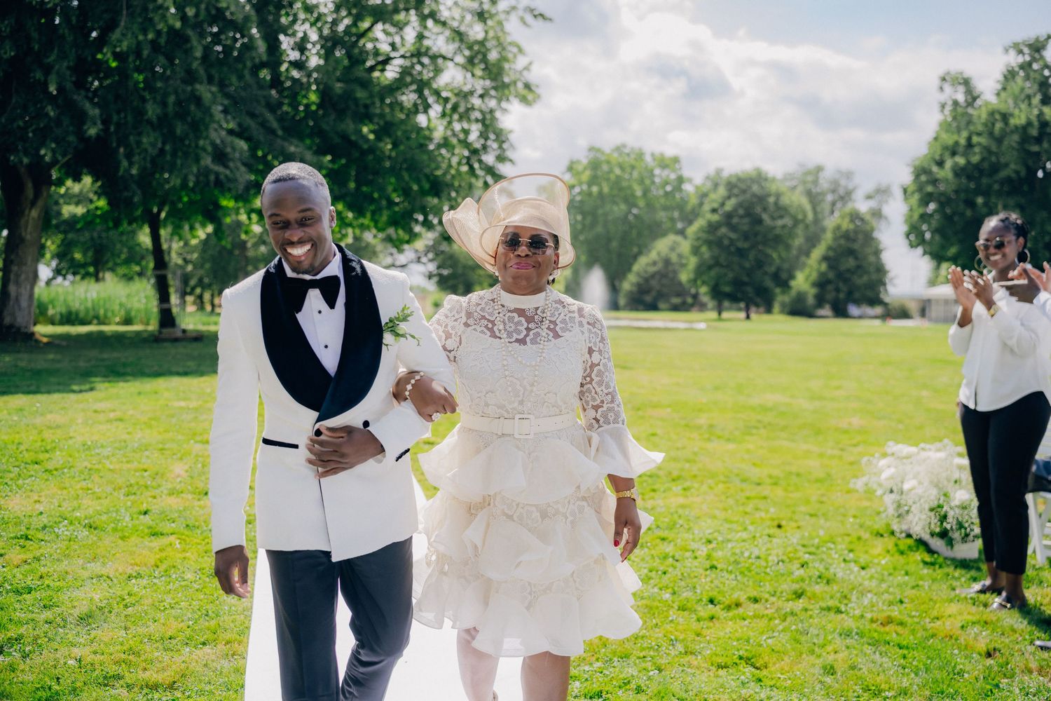 A couple in wedding attire walks together across a sunny green lawn during their outdoor celebration.