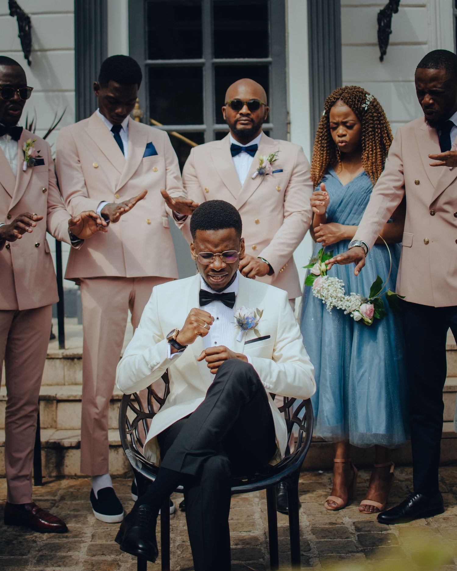 A wedding party poses in matching pink suits and formal attire on stone steps in front of a white building.
