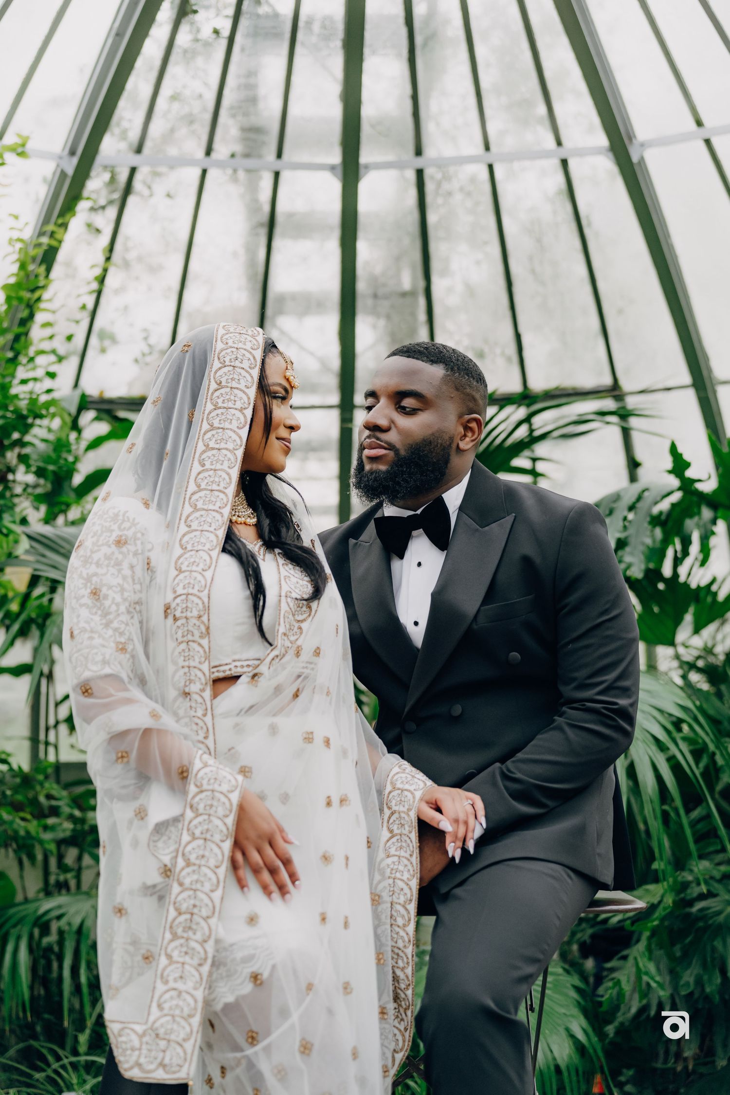A couple shares an intimate moment in a greenhouse wearing elegant wedding attire with lush greenery surrounding them.
