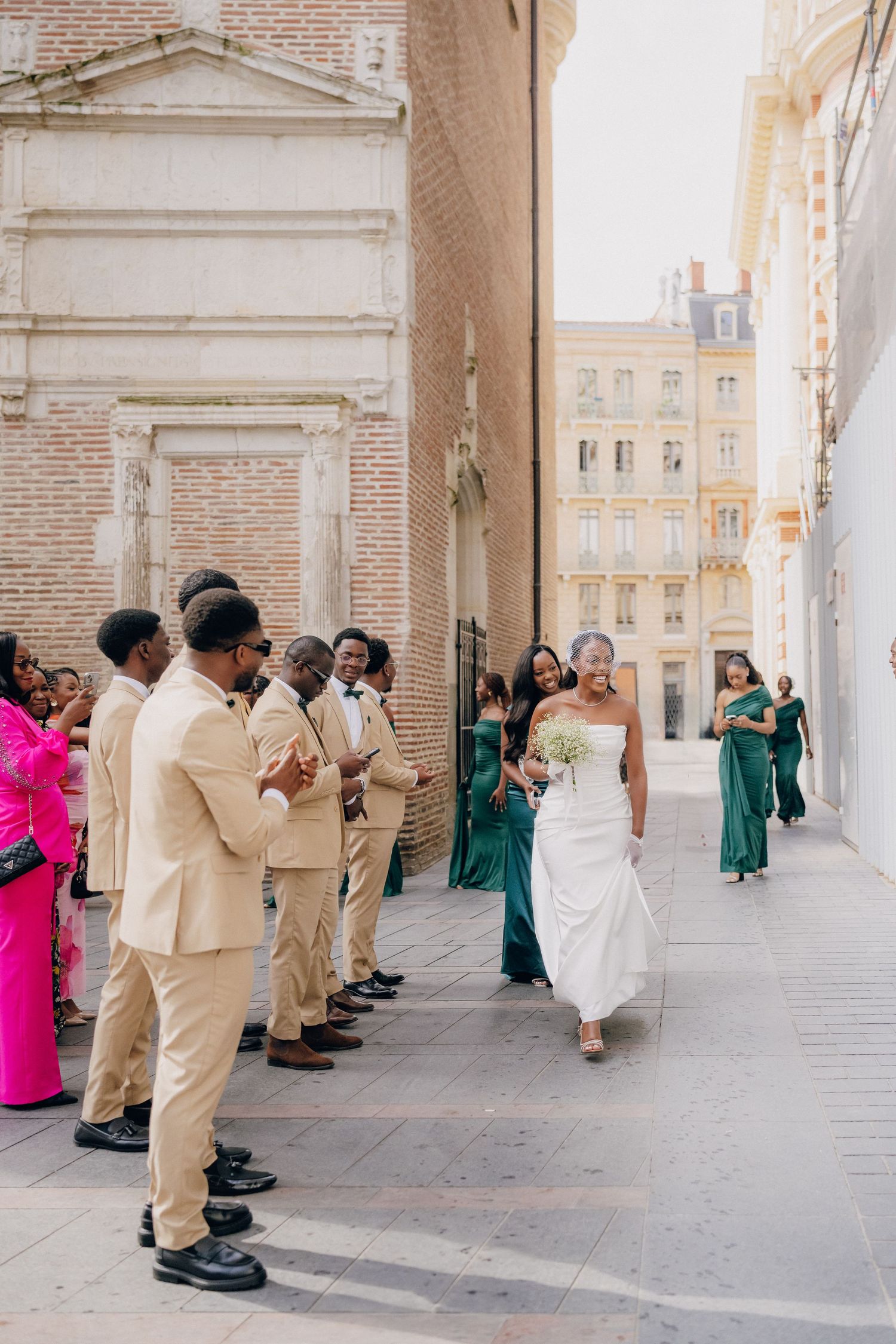 Wedding party poses for photos in elegant archway with bridesmaids in green dresses and groomsmen in tan suits.
