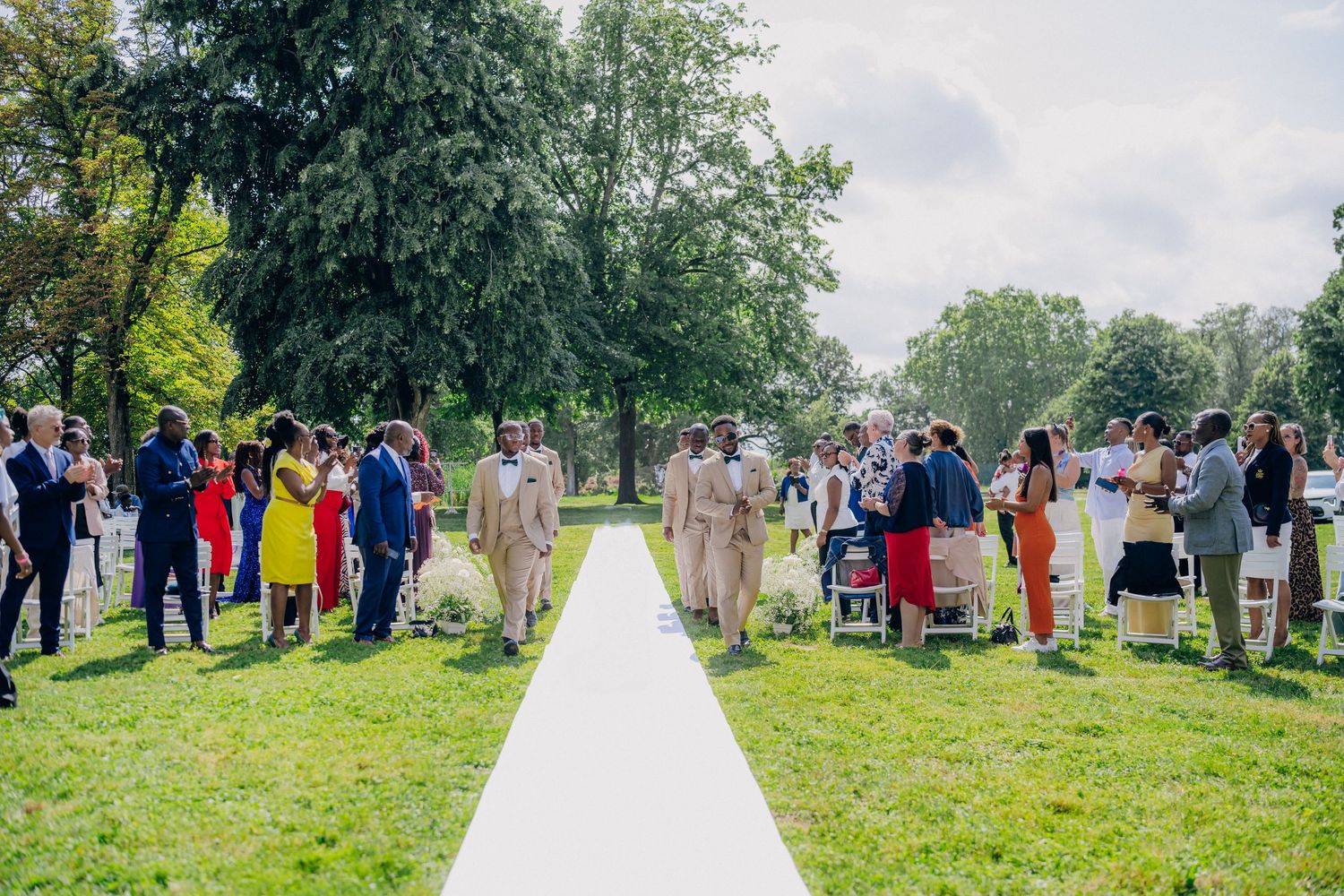 A wedding ceremony in a lush green outdoor setting with guests gathered along a white aisle runner.