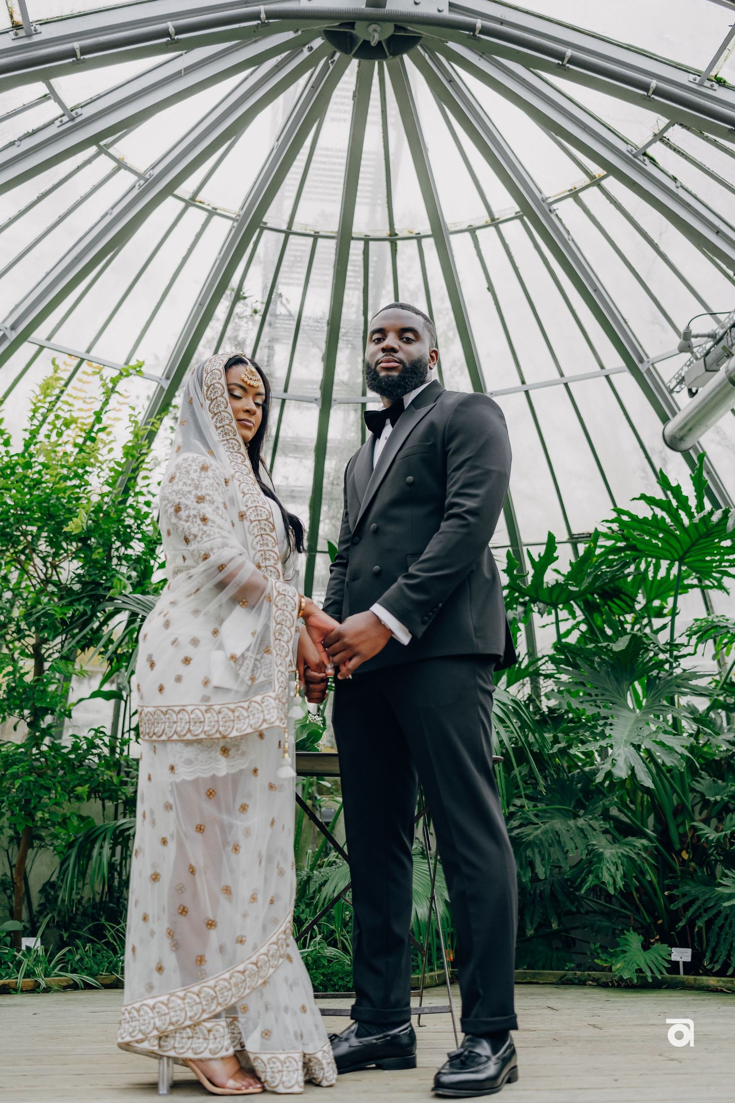 Newlywed couple stands together in a sunlit greenhouse dome with tropical foliage.