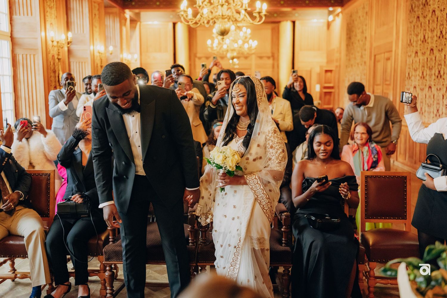 A wedding celebration in a grand room with guests seated and a chandelier overhead.