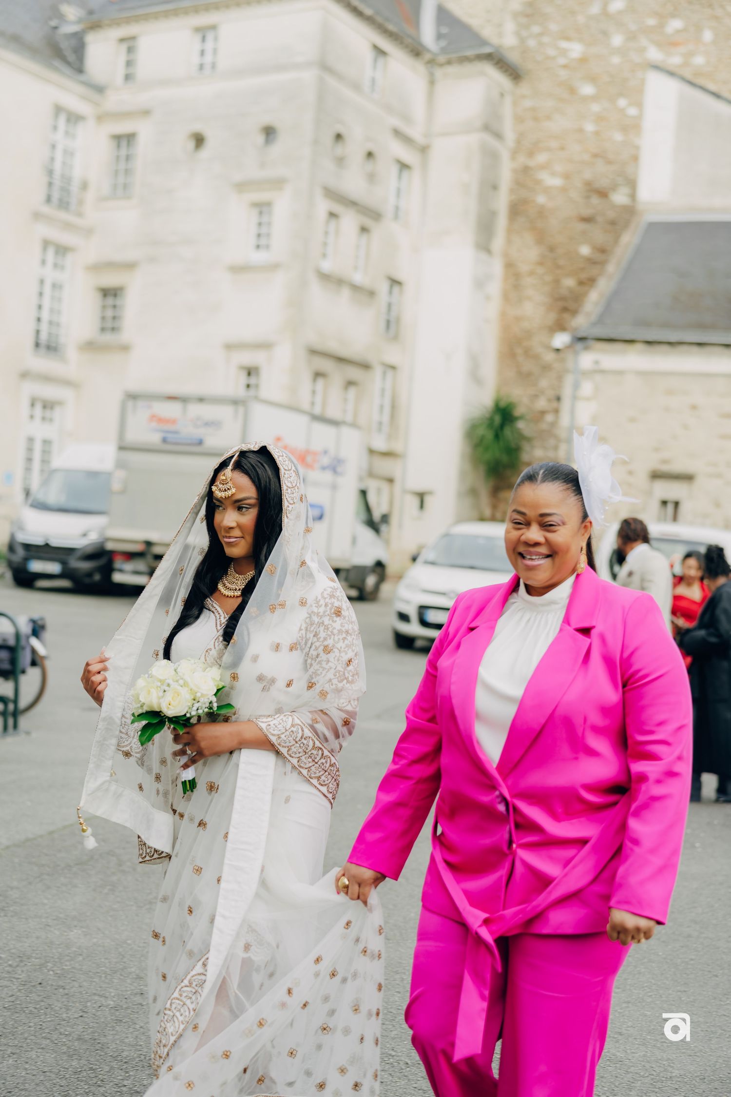 A celebration outside a historic building with a bride in white dress and companion in bright pink suit.