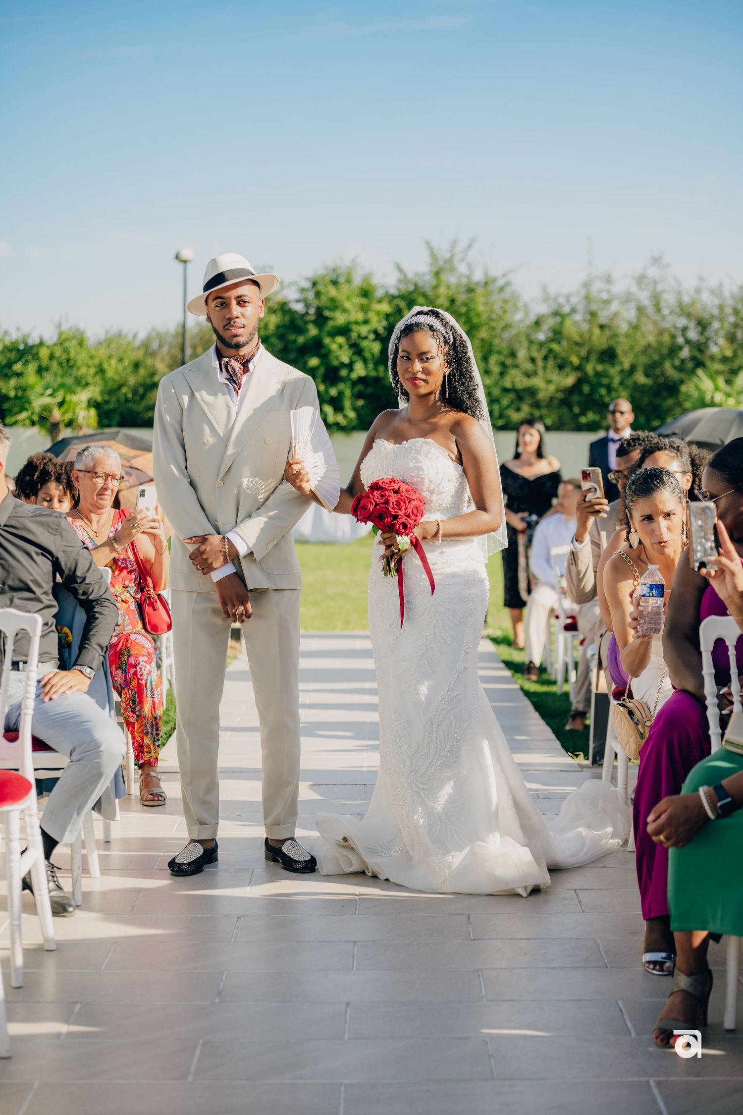 A couple walking down the outdoor wedding aisle while guests watch from white chairs on a sunny day.