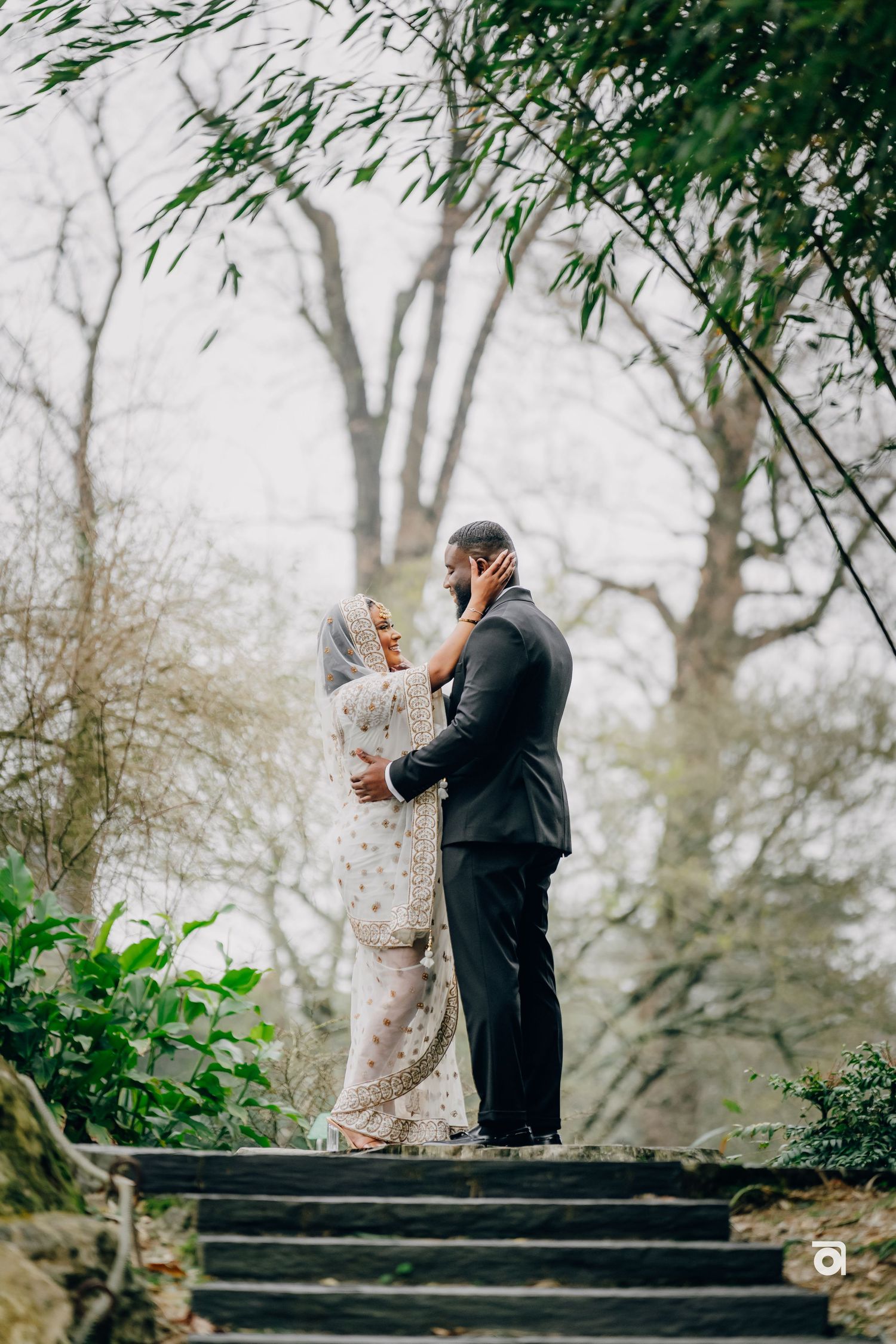 A couple in wedding attire share a tender moment standing on misty garden steps surrounded by greenery and trees.