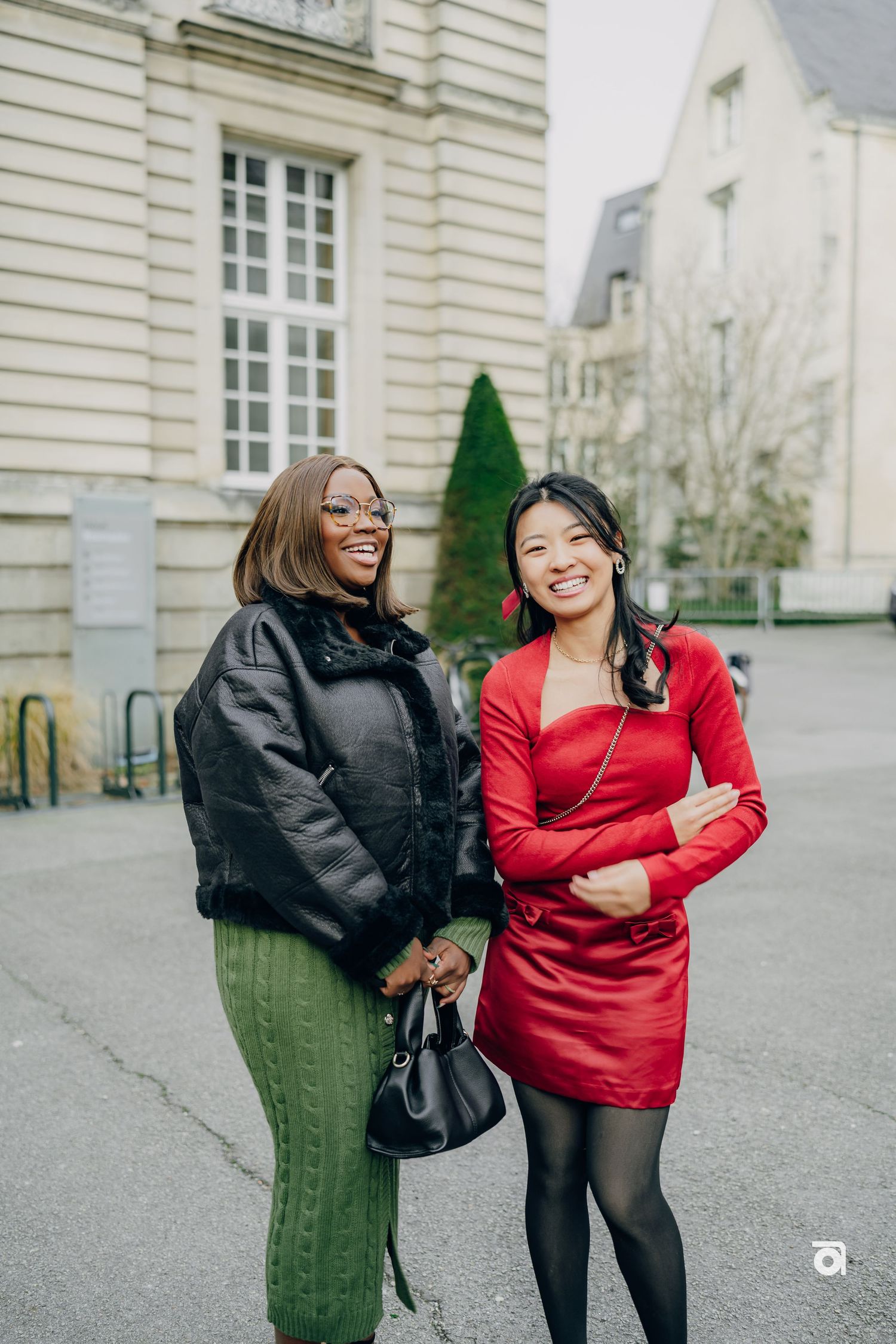 Two friends laughing together on a street in front of classic European architecture, one in a red dress and one in black coat.