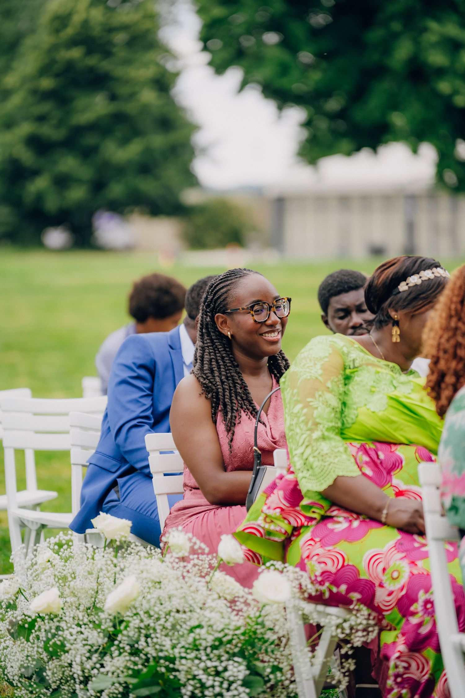 Wedding guests seated in colorful attire among floral arrangements and white chairs at an outdoor ceremony.