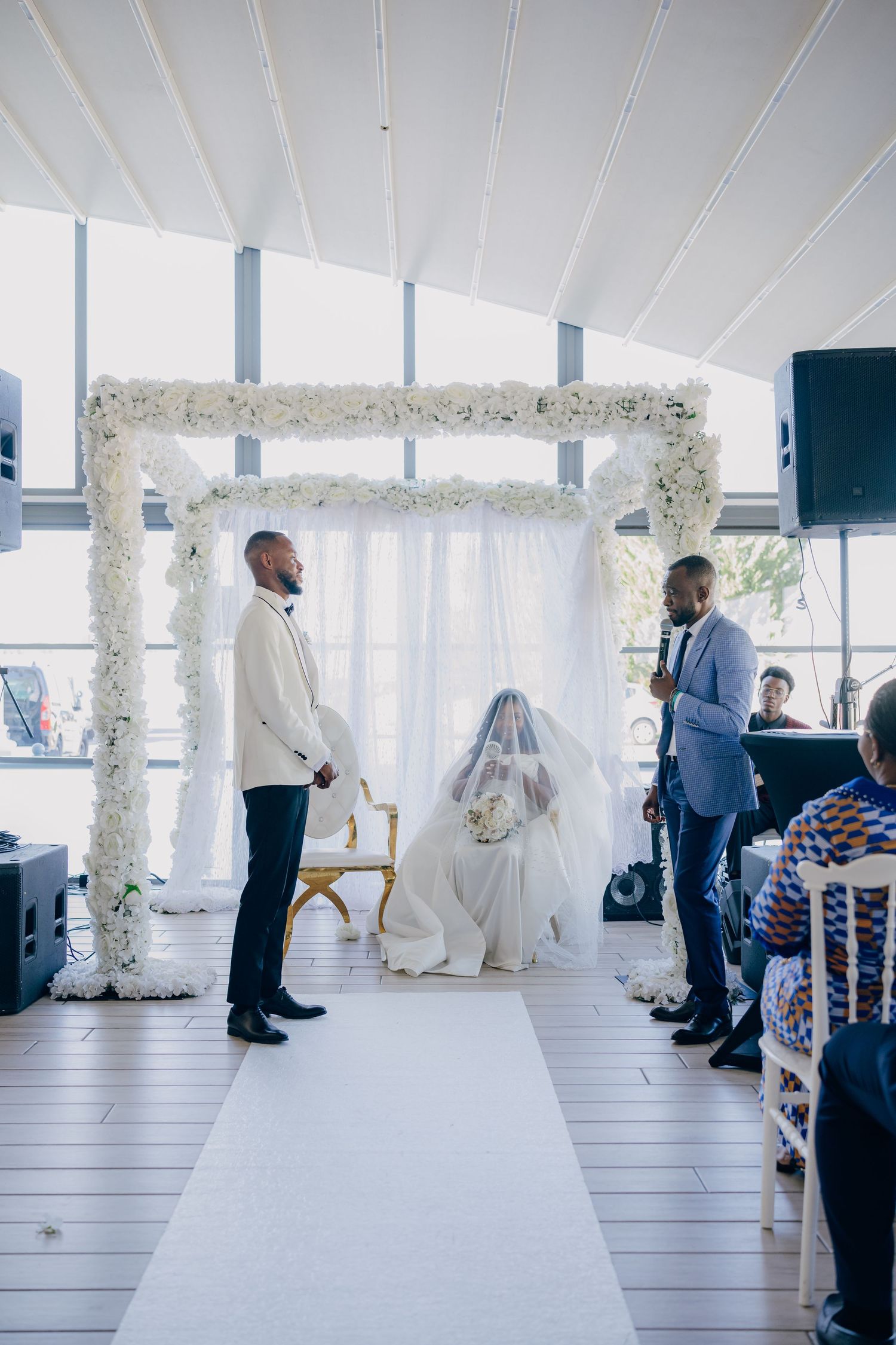 Wedding ceremony in progress in a bright modern venue with white floral archways and seated guests.