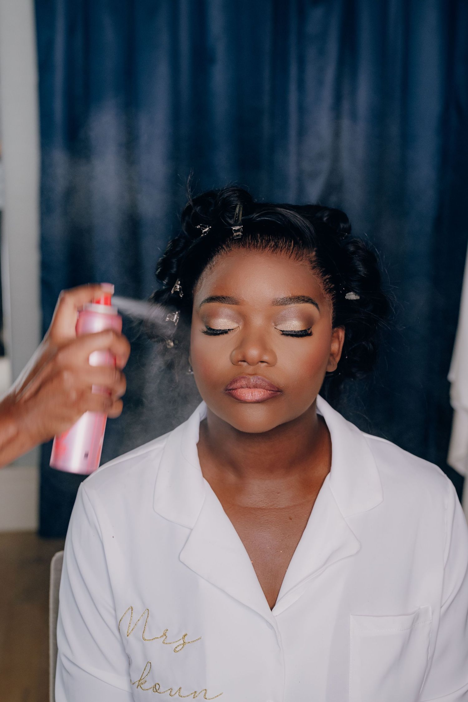 A makeup artist sprays setting spray on a model wearing a white robe during a beauty session.