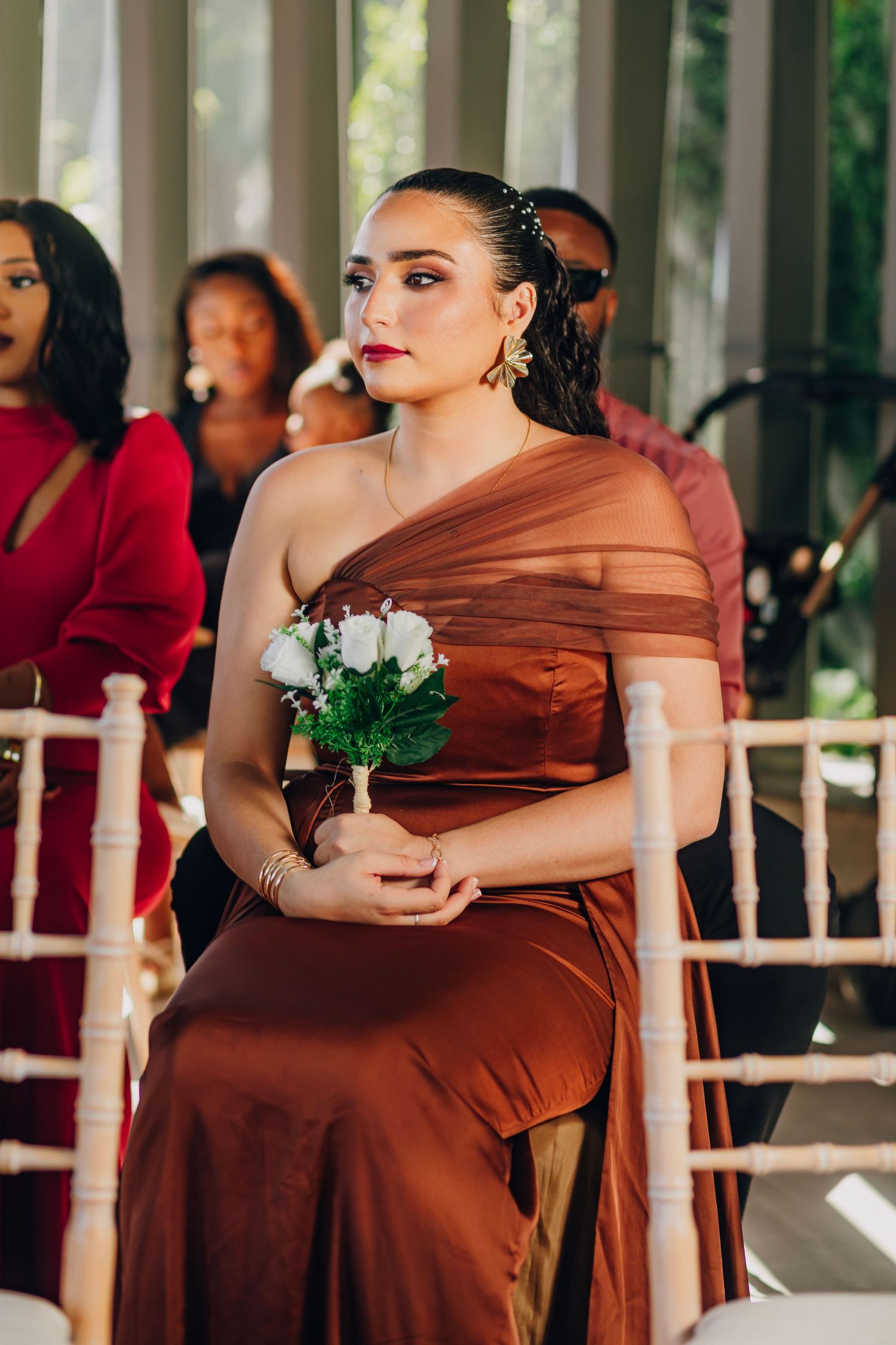 A bridesmaid in a brown one-shoulder gown holds a small white bouquet during a wedding ceremony.