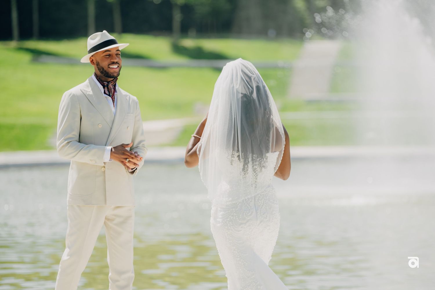 A bride in a white dress and a groom in a cream suit share a moment by a fountain on their wedding day.