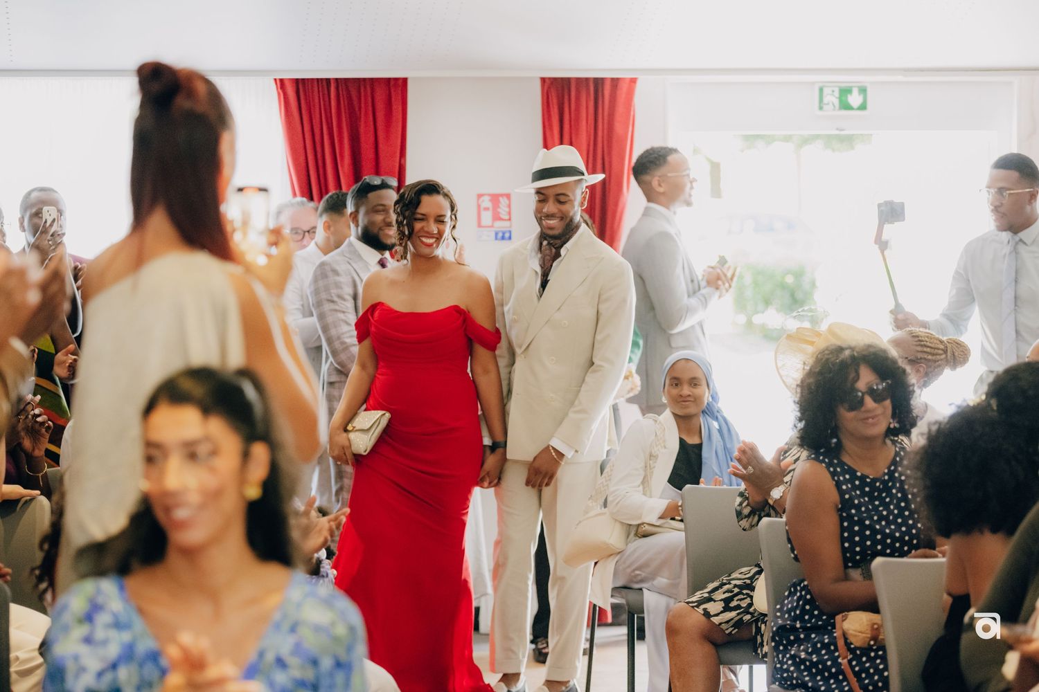 Wedding guests mingle at an indoor reception venue decorated with red curtains and bright lighting.