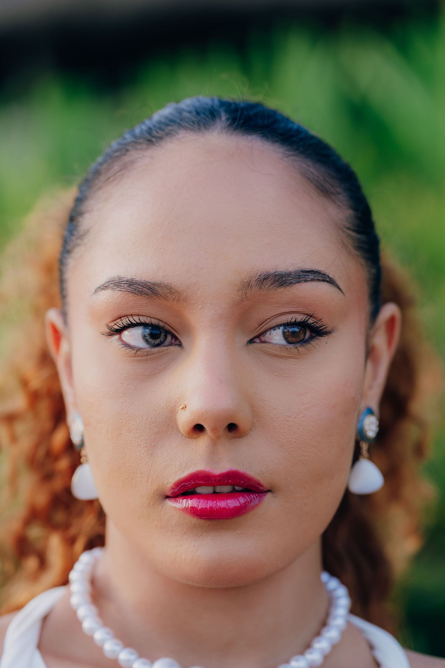 Close-up portrait of a young woman with red lips, nose ring, pearl earrings, and necklace against green background.