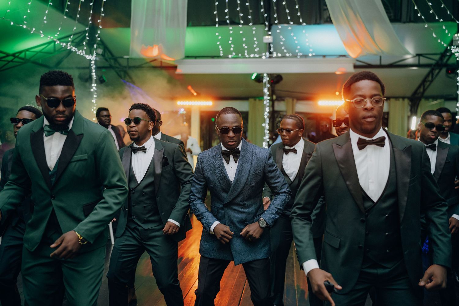 A group of groomsmen in black tuxedos and bow ties pose together under decorative string lights at a wedding reception.