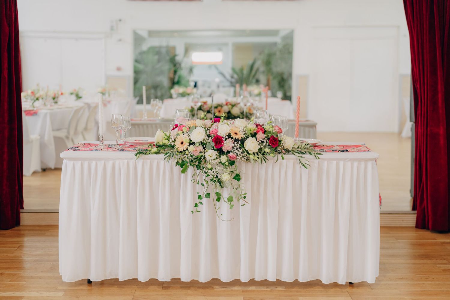 Elegant wedding reception head table with white linens and pink floral centerpiece arrangement in a bright indoor venue.
