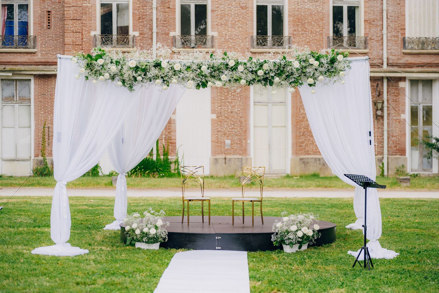 Elegant outdoor wedding ceremony setup with white draping and floral arrangements against a brick building.