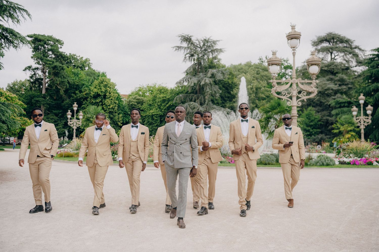 Wedding party members in beige and gray attire walking in line formation through a garden setting.