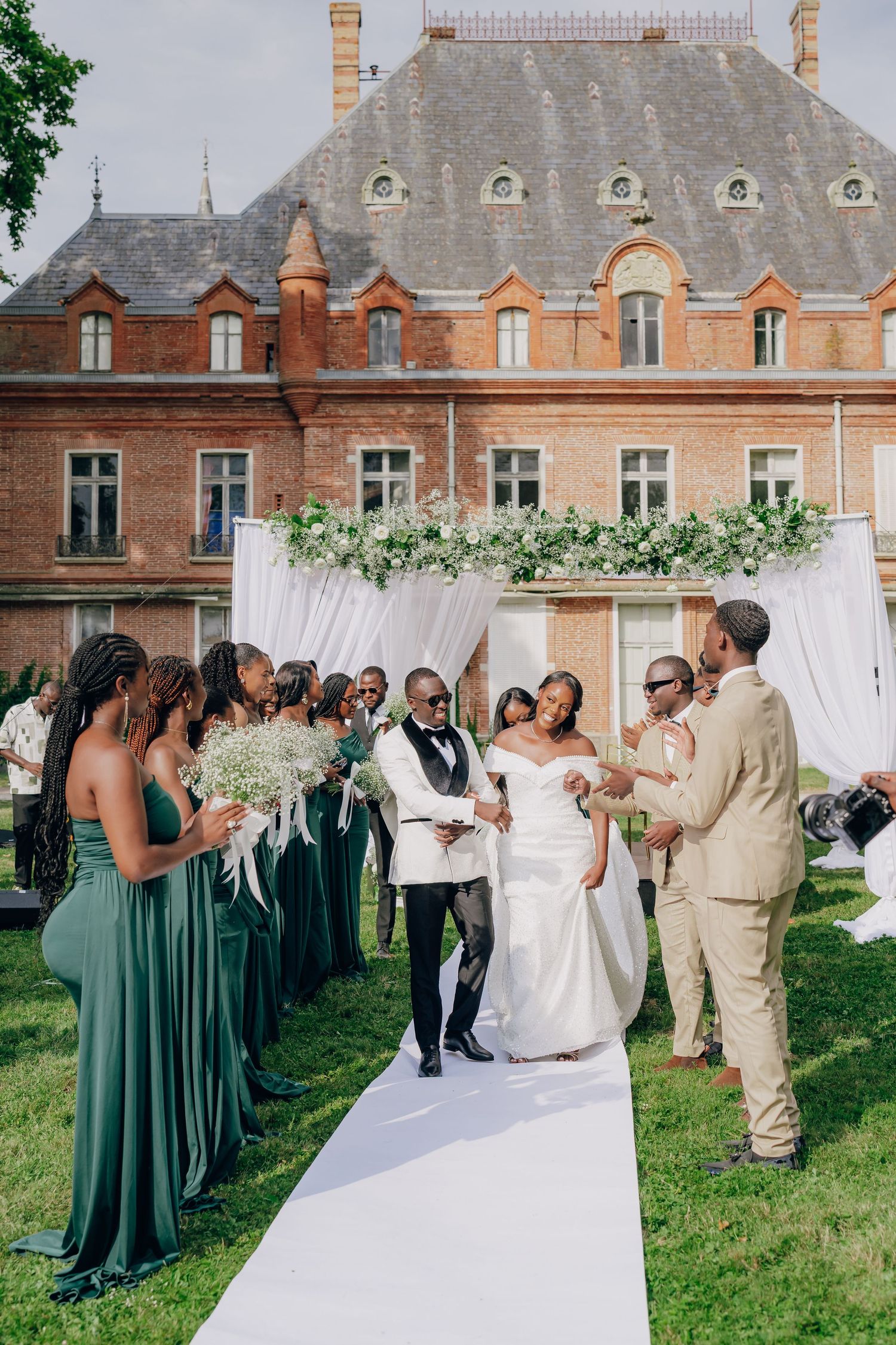 Wedding party in green dresses and beige suits celebrates on a manicured lawn with historic brick buildings in background.