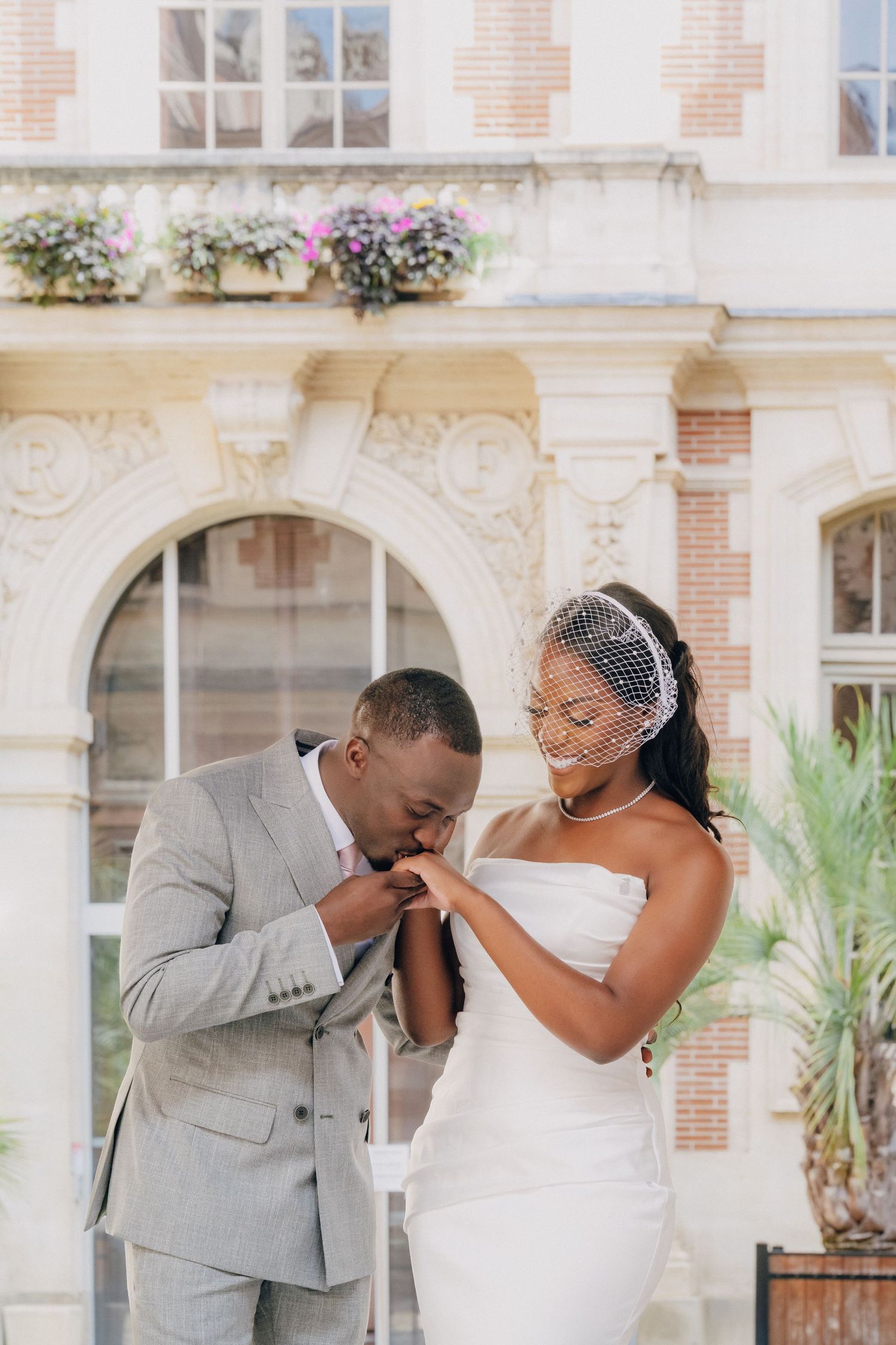 Romantic wedding moment captured in front of elegant brick building with ornate architecture and flower boxes.
