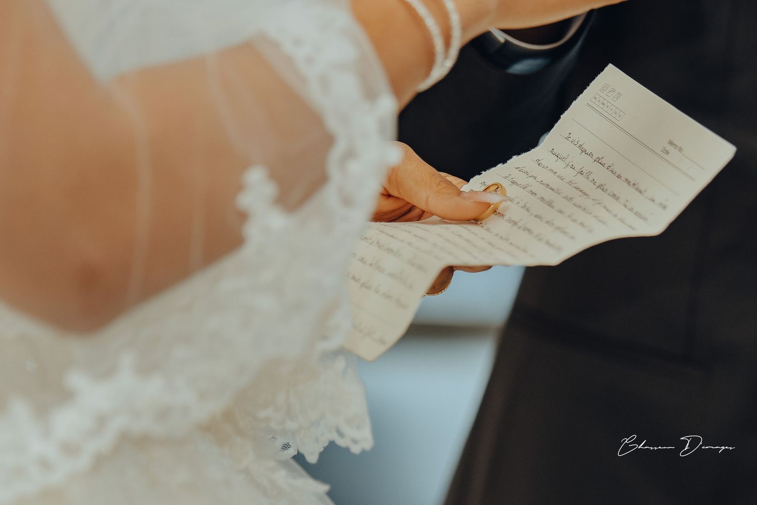 Close-up shot of hands holding wedding vows written on paper with lace dress detail in frame.