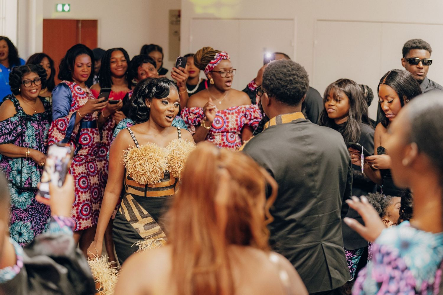 Wedding guests wearing colorful traditional African print dresses gather to celebrate at an indoor celebration.