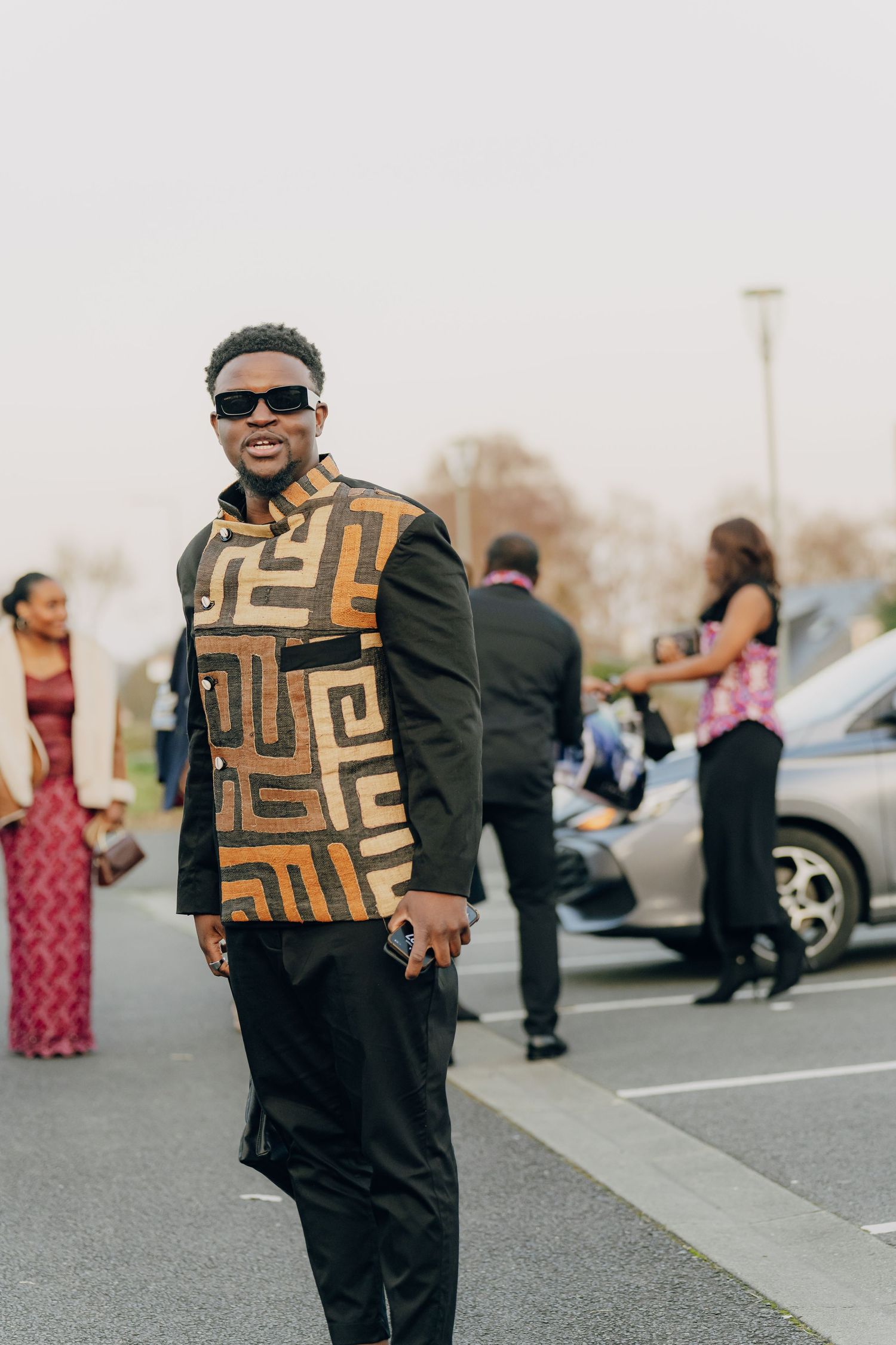 Stylish person in geometric patterned jacket and sunglasses poses in a parking lot with people visible in background.