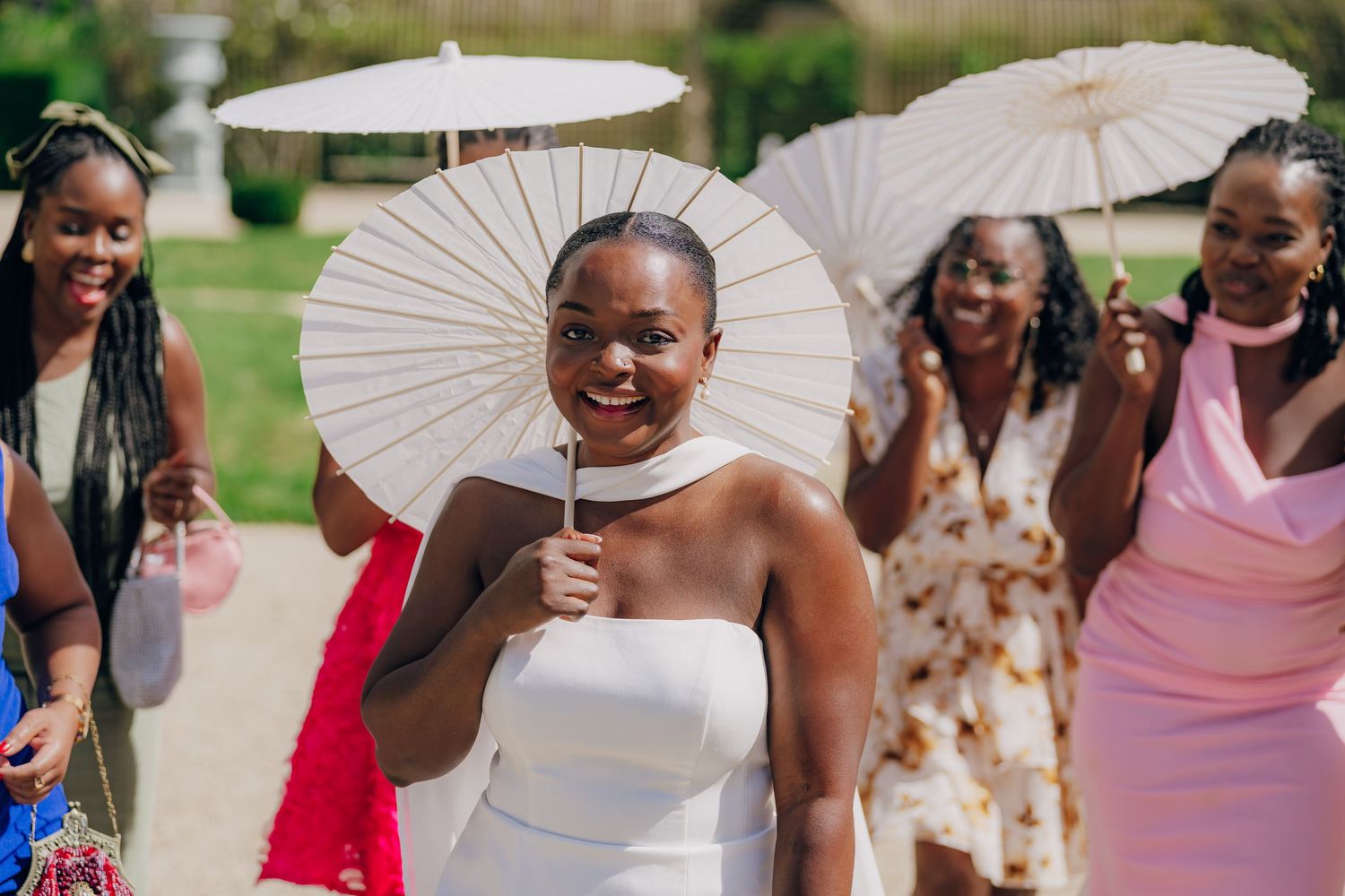Wedding party members laugh and hold white parasols while walking together outdoors on a sunny day.