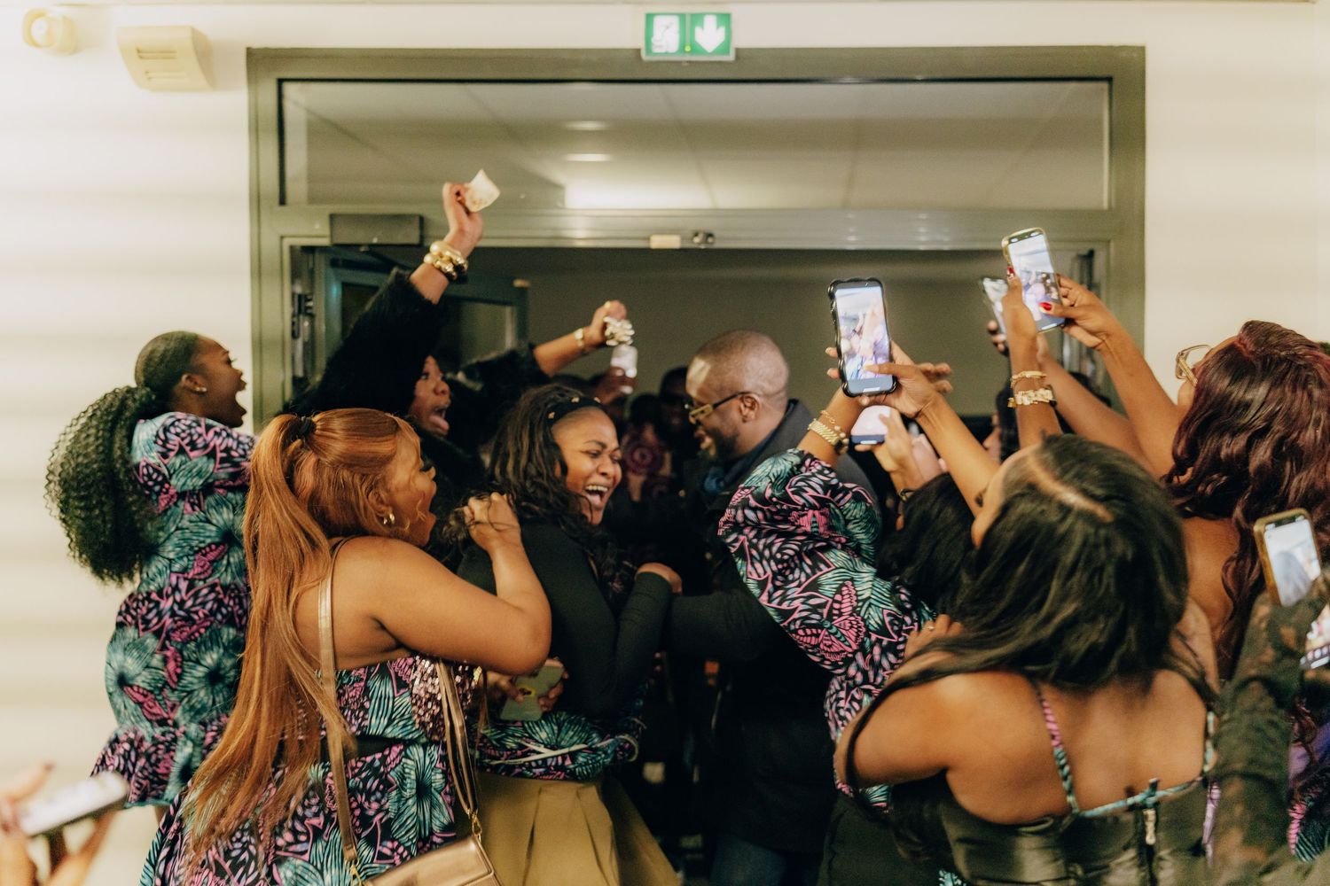 A group of people dressed in floral prints celebrate with raised phones and drinks on a dance floor.