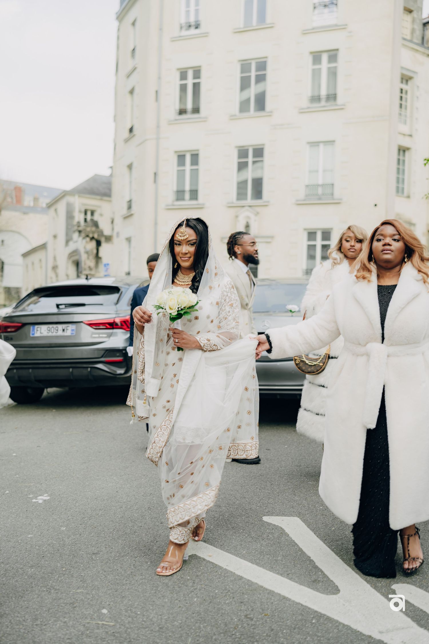 A group walking on a city street in elegant white outfits near a parked luxury car.