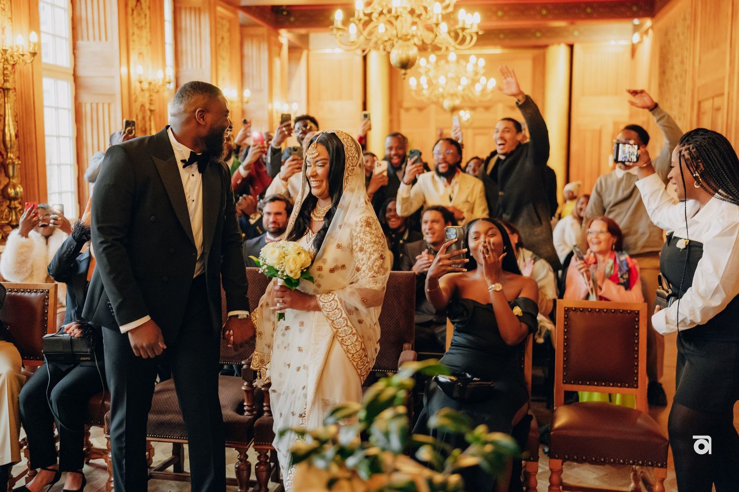 Wedding guests celebrate in an elegant room with wooden details and chandeliers.