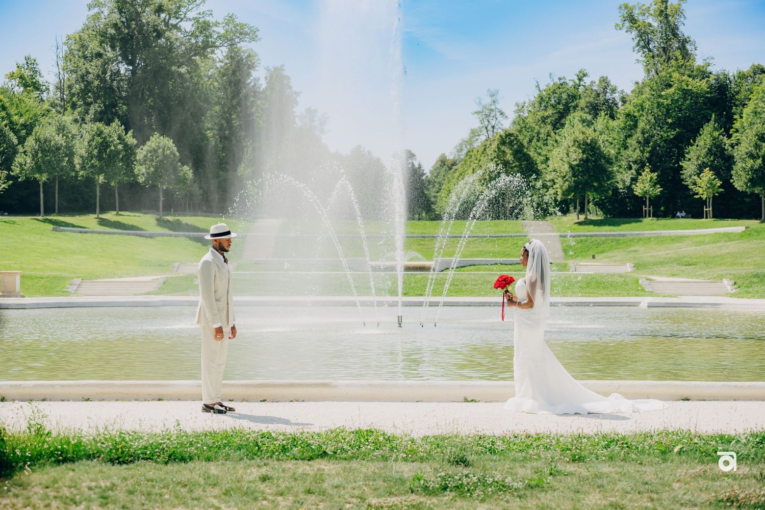 A wedding couple stands on opposite sides of a fountain spraying water in a sunny park setting.