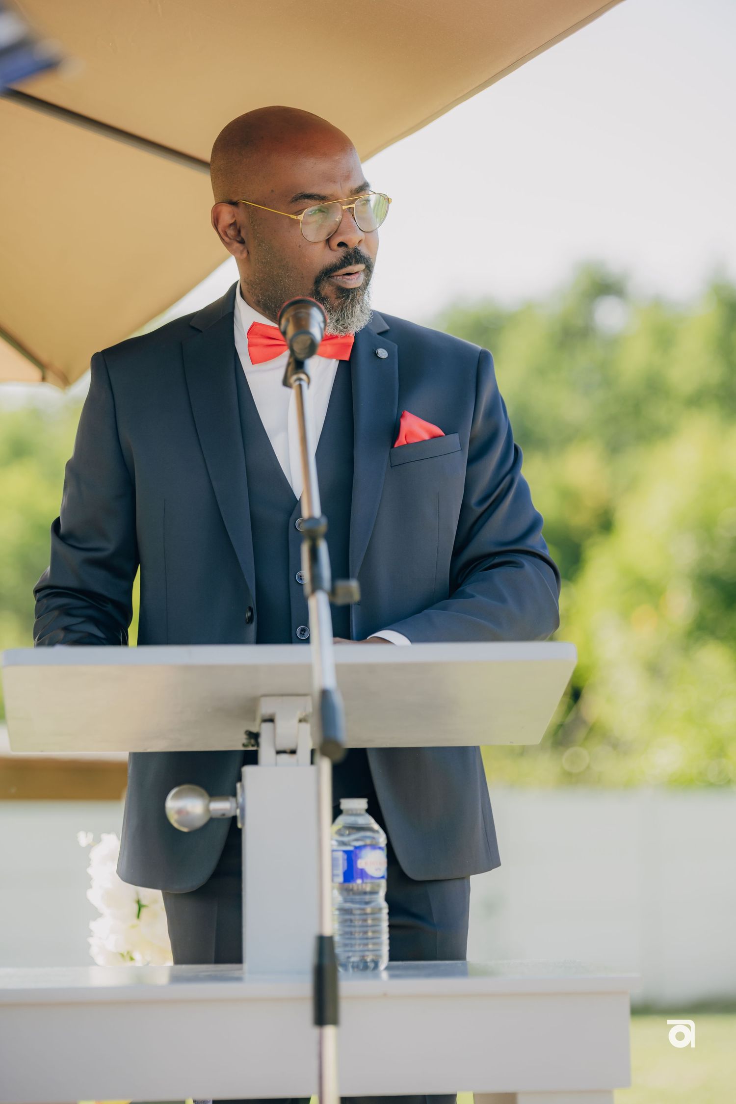 A speaker in a navy suit and red bowtie stands at a podium delivering remarks during an outdoor event.