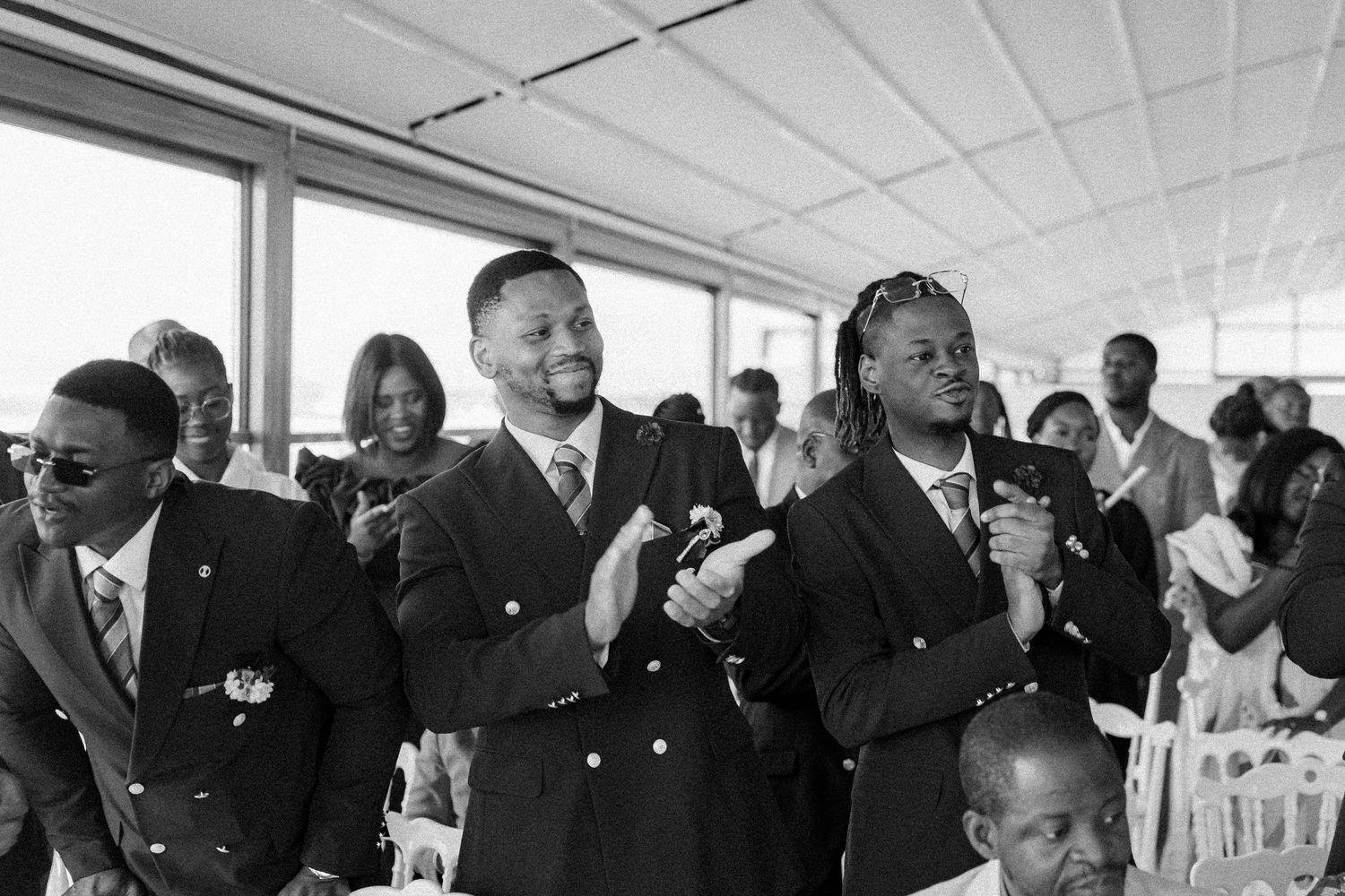 Wedding guests in formal black attire react emotionally during a ceremony in a bright indoor venue.