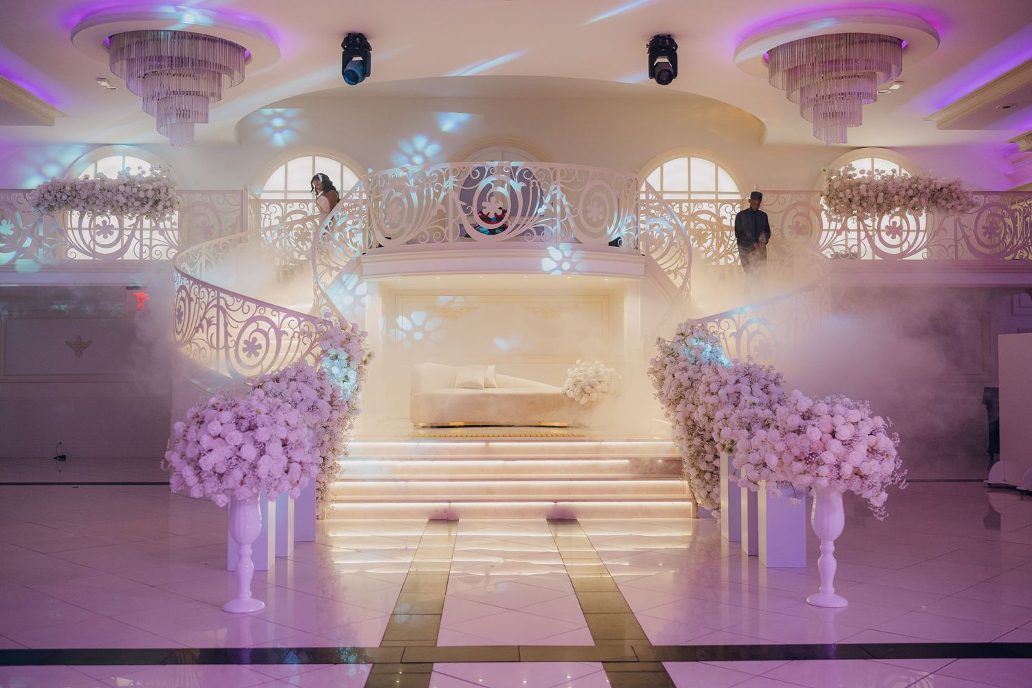 Elegant wedding venue setup with purple lighting, crystal chandeliers, and floral arrangements on pedestals.