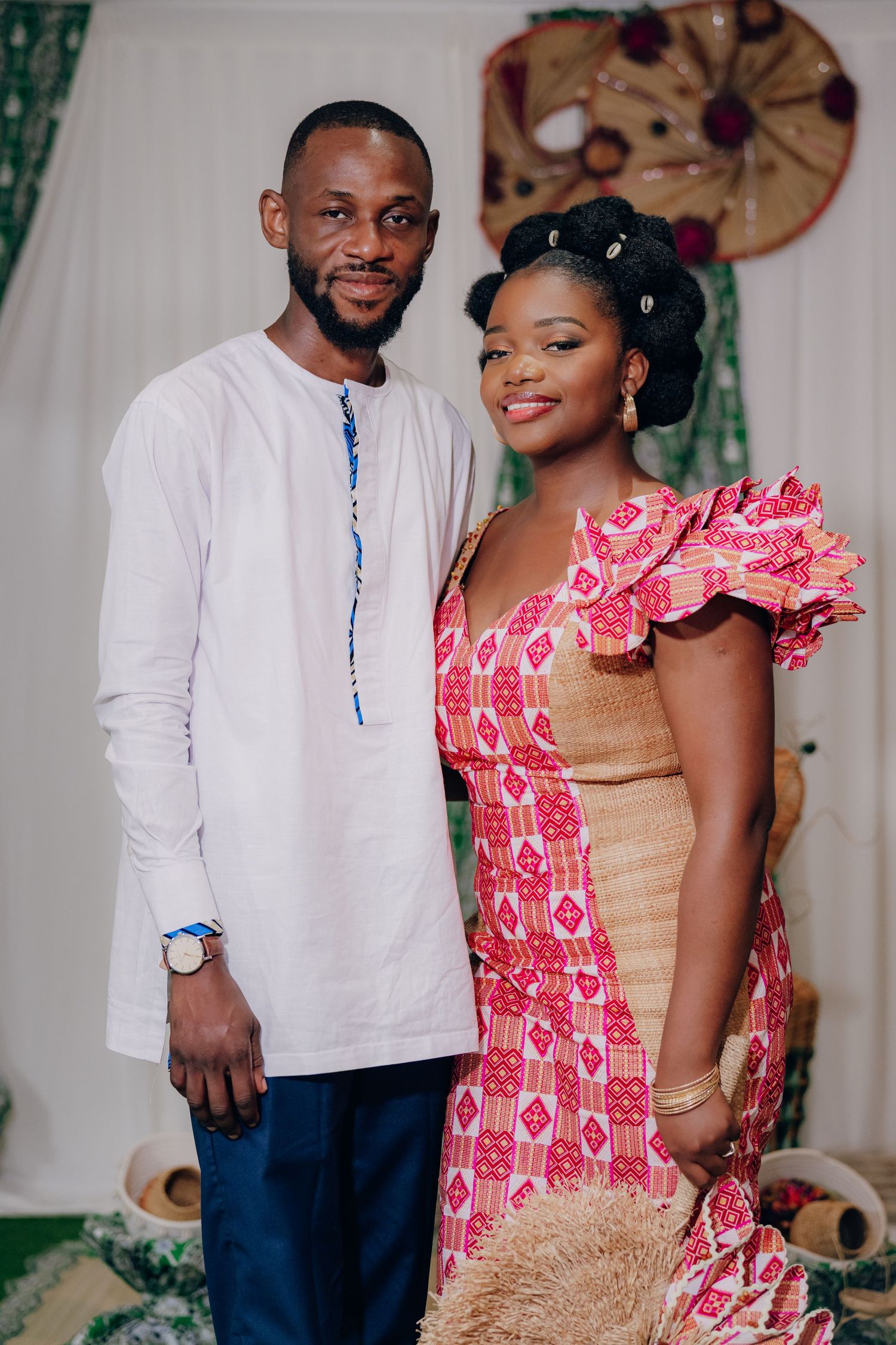 A couple poses together in traditional attire, with the bride wearing a pink patterned dress and the groom in white.