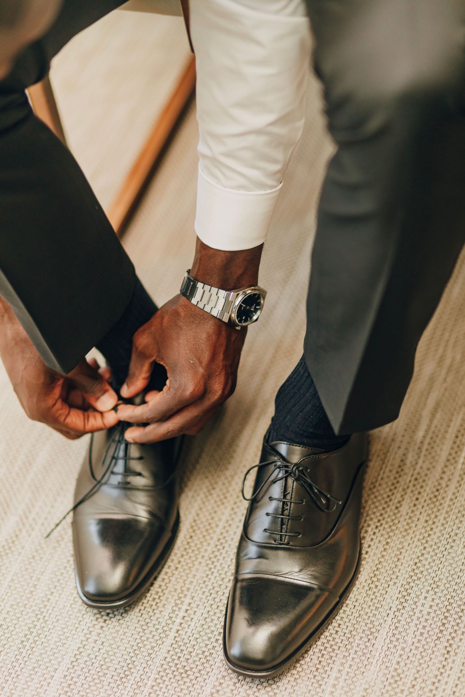 Close-up of hands tying dress shoe laces with a silver watch visible on the wrist.