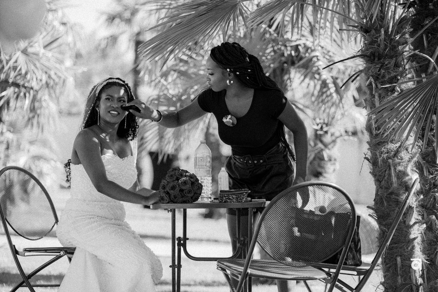 Two people share a candid black and white moment seated at a small table surrounded by tropical foliage.