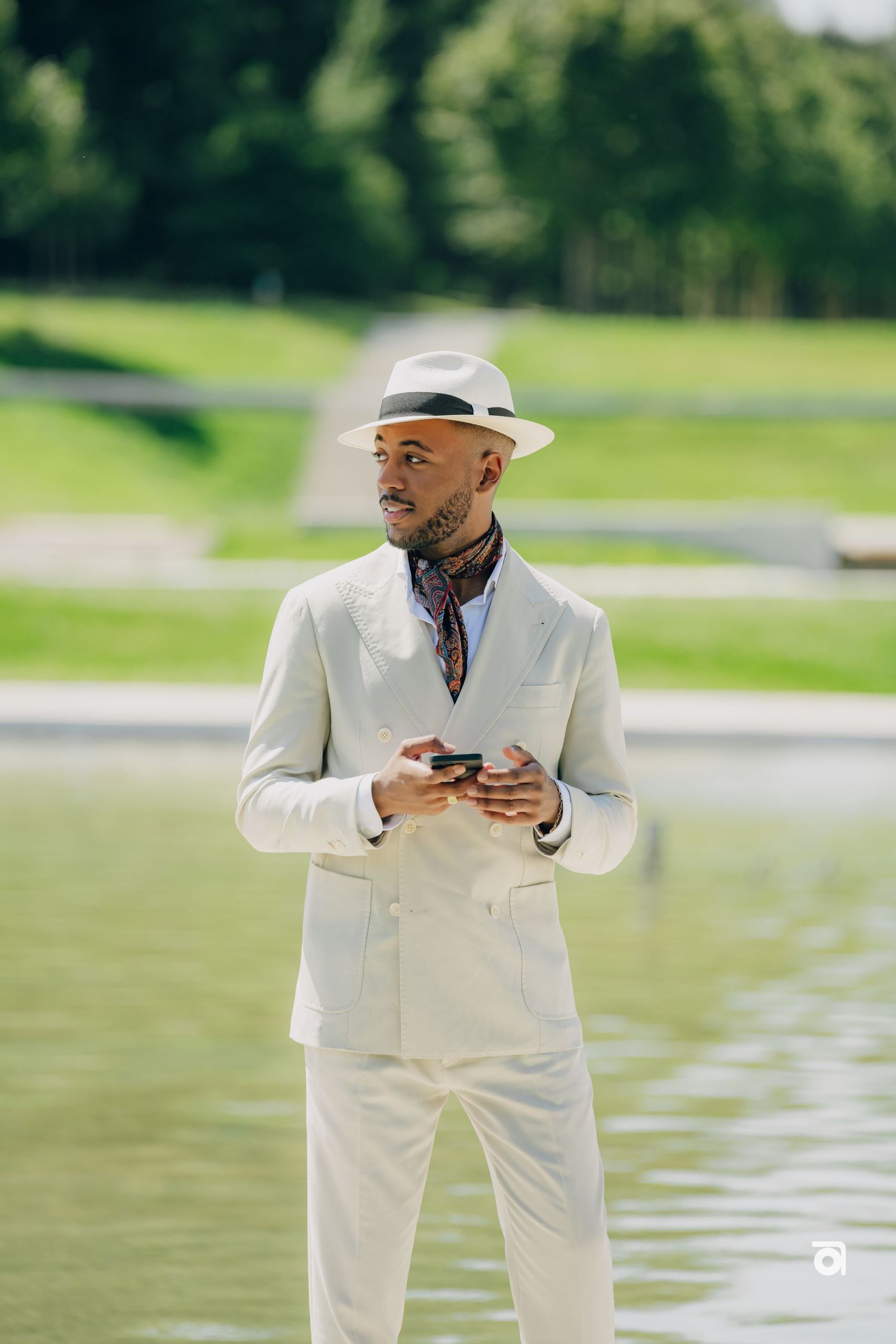 Stylishly dressed person in an all-white suit and fedora checks phone while standing near a serene lake on a sunny day.