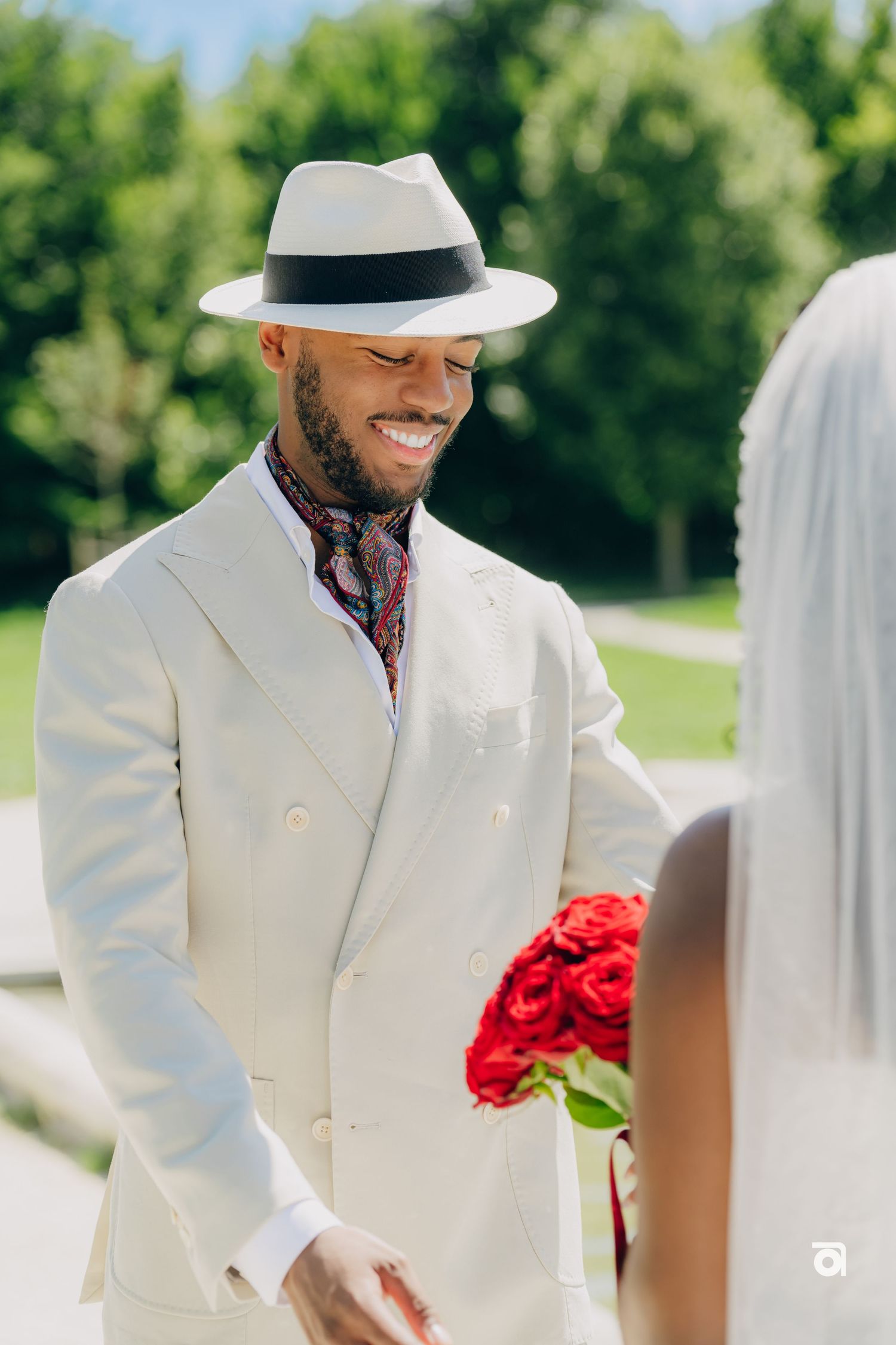 Groom in white suit and panama hat shares a joyful moment with bride holding red roses.