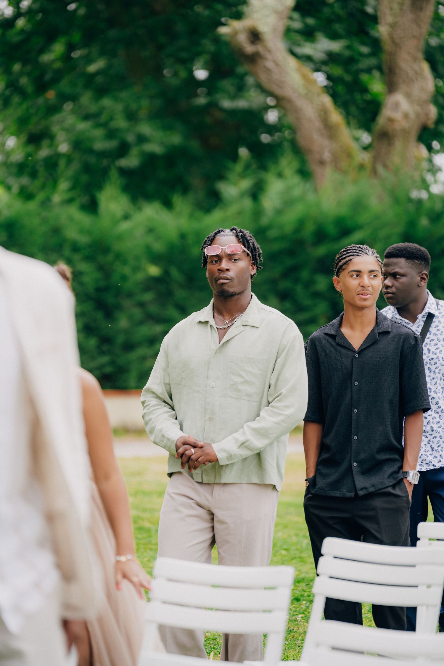 Wedding guests in formal attire gather outdoors under green trees during a summer celebration.