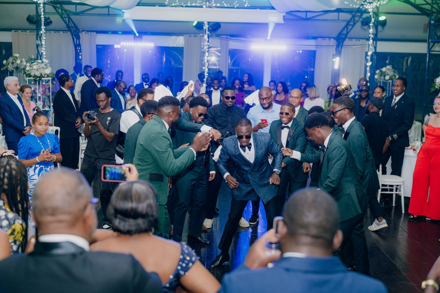 Wedding party members dancing and celebrating in formal attire under blue lighting at an indoor venue.