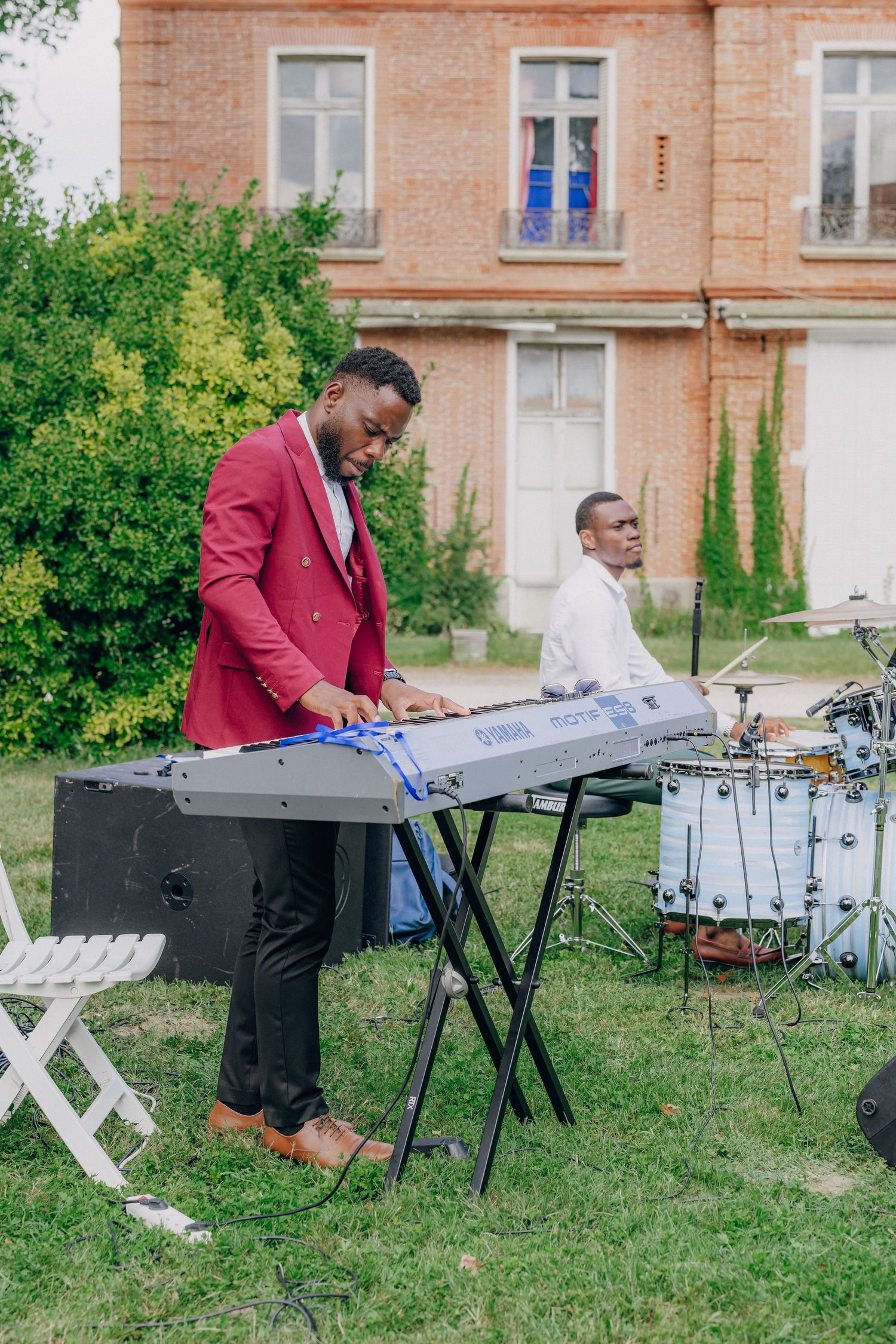 Musician in burgundy jacket playing keyboard outdoors near a brick building.