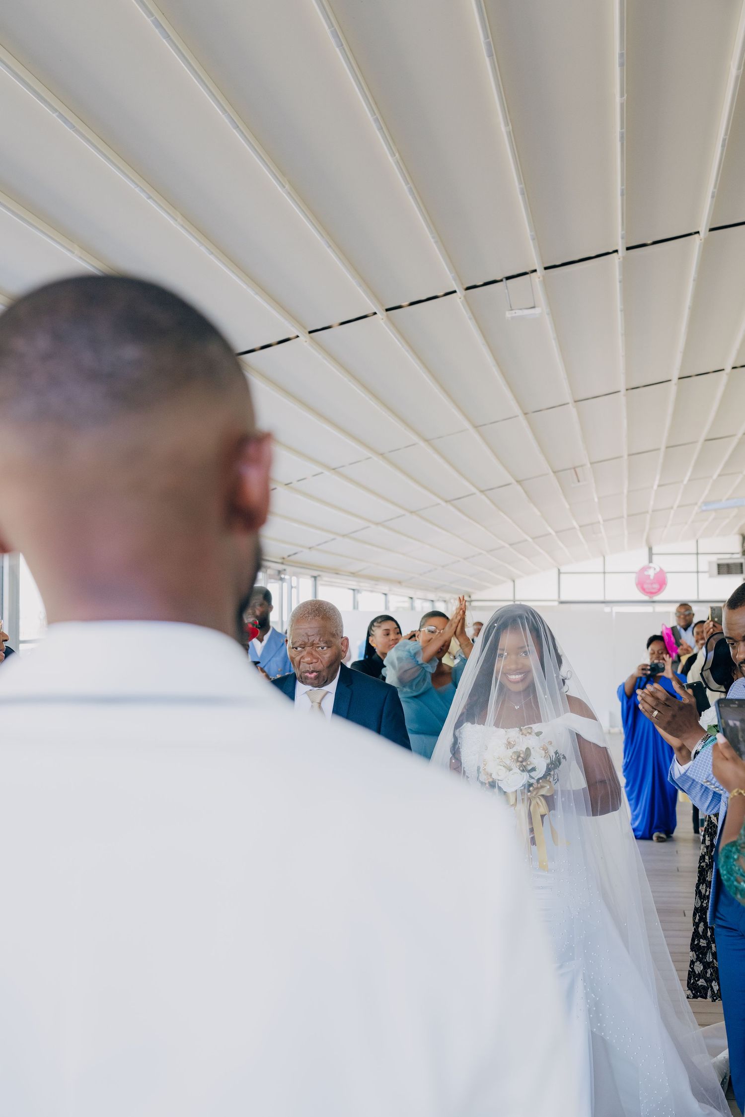 A wedding ceremony scene with guests in formal attire gathered in an indoor venue with white decor.
