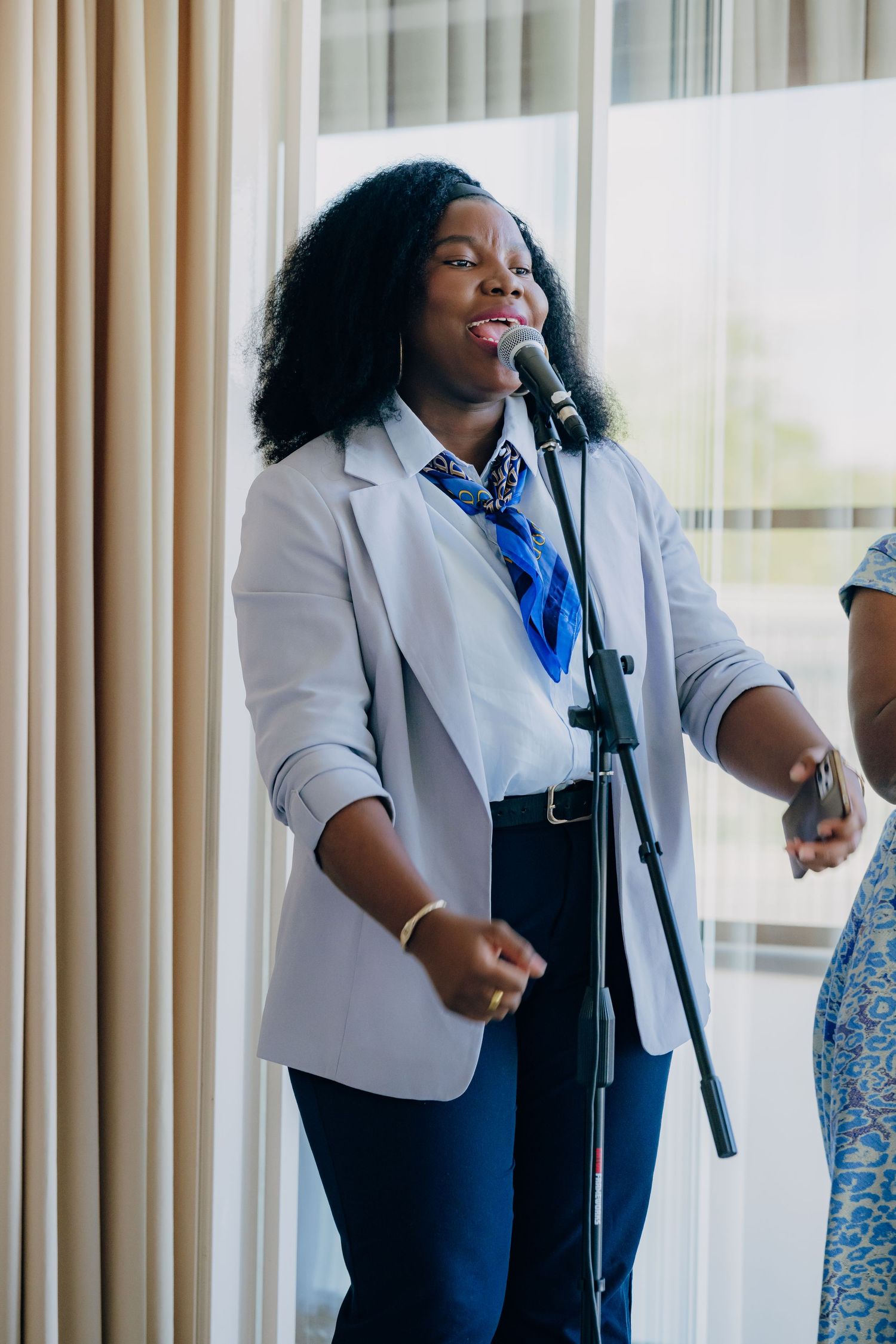 A performer in a gray blazer and blue tie sings into a microphone near curtained windows.