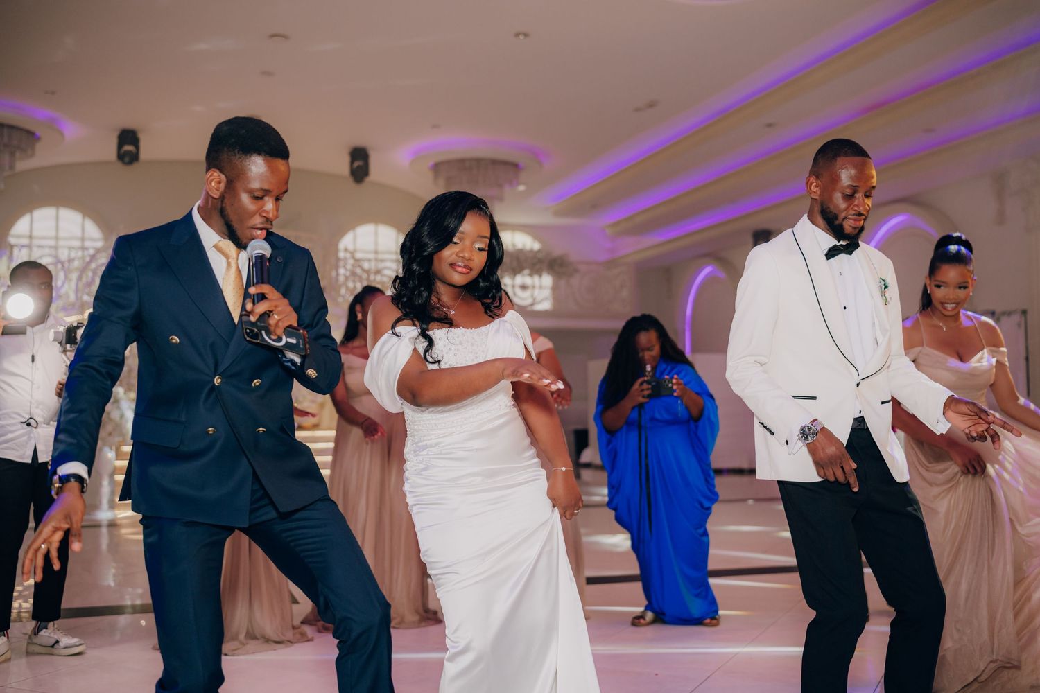 Wedding guests dancing and celebrating at a reception with purple lighting and white decor.