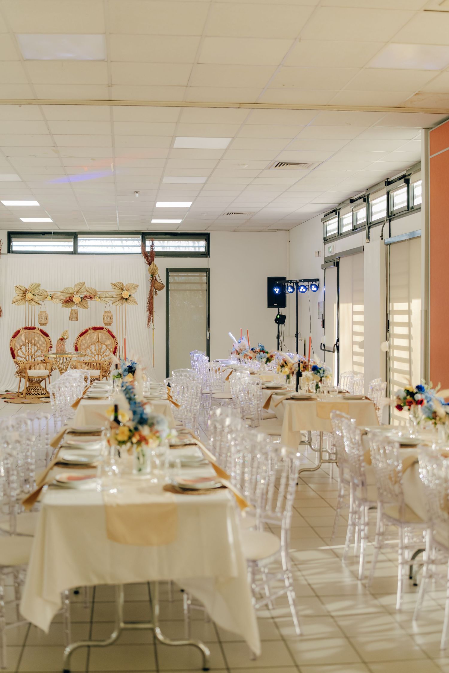 Modern wedding reception setup in a banquet hall with elegant white tables, clear chairs and decorative floral centerpieces.