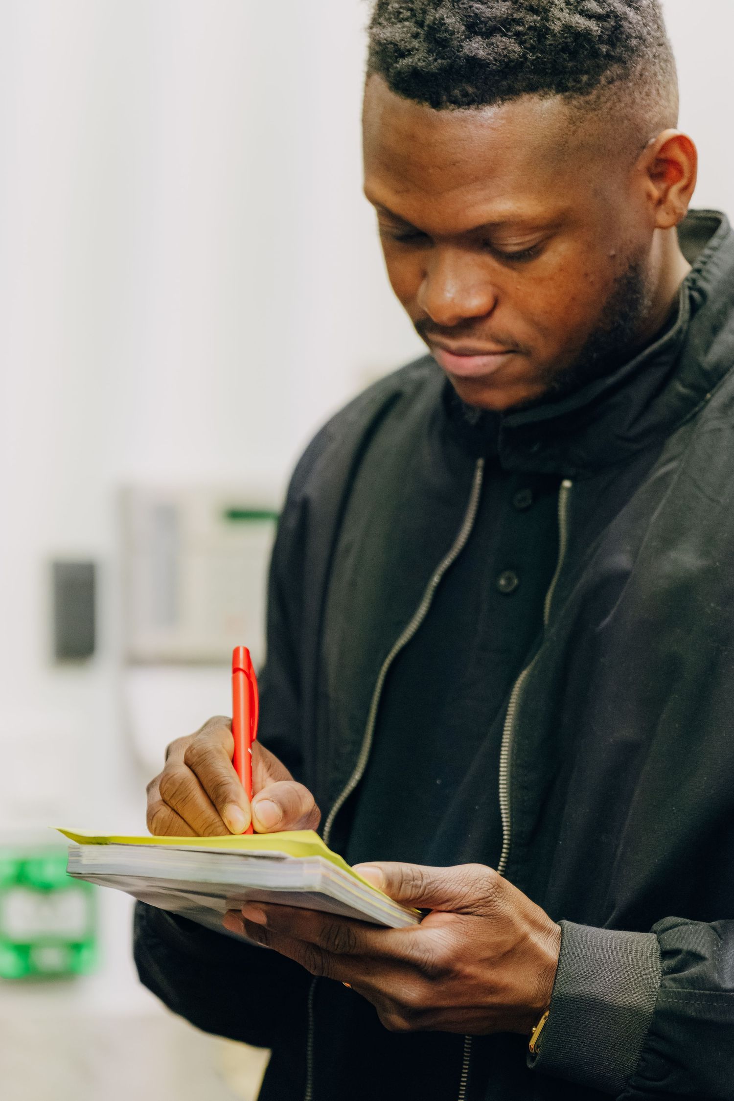 Person in black jacket writes with red pen in a notebook while standing indoors.