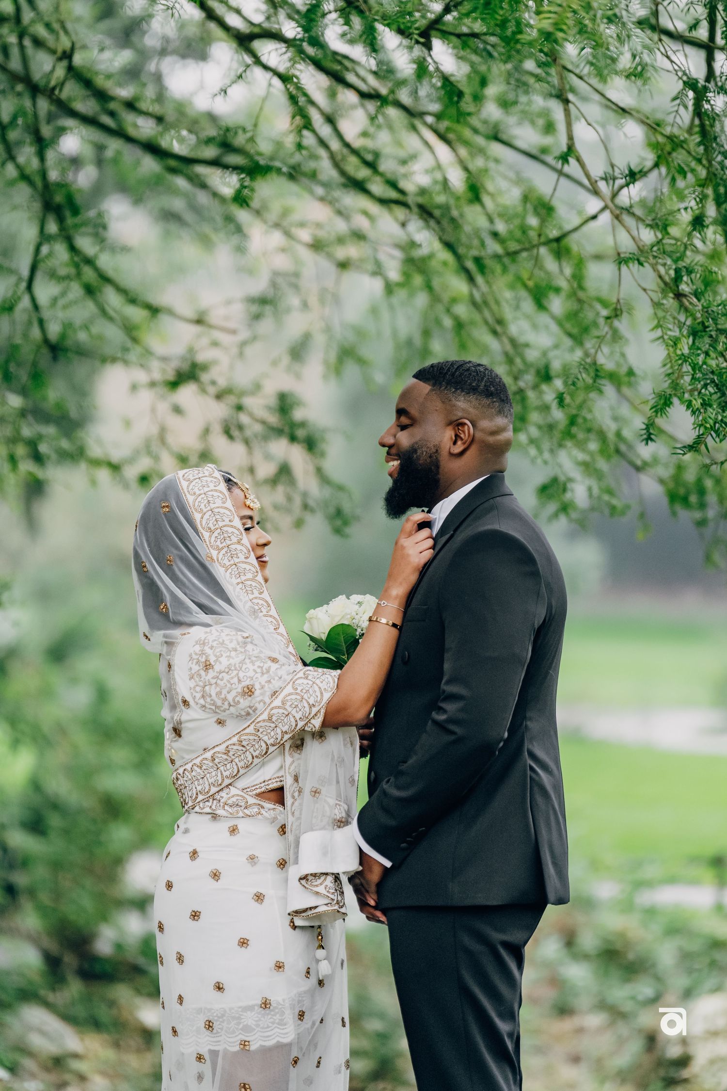 A couple shares an intimate moment under flowering trees, with the bride wearing an ornate white and gold dress and veil.